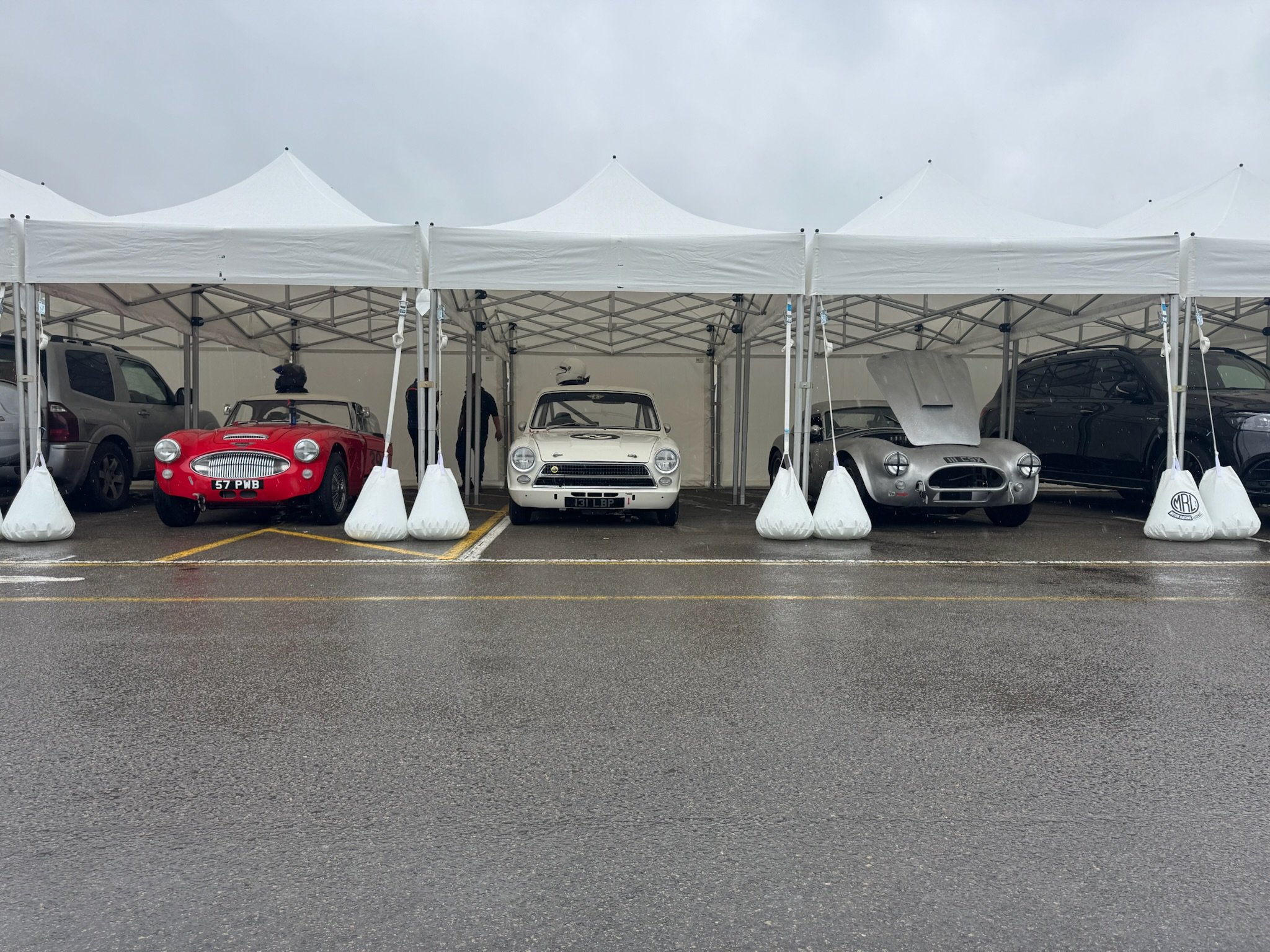 A white canopy tent at a car show, displaying five vintage cars, including a red sports car, a white car with a hood, and a silver car with a sleek design, parked on a paved surface on a cloudy day.