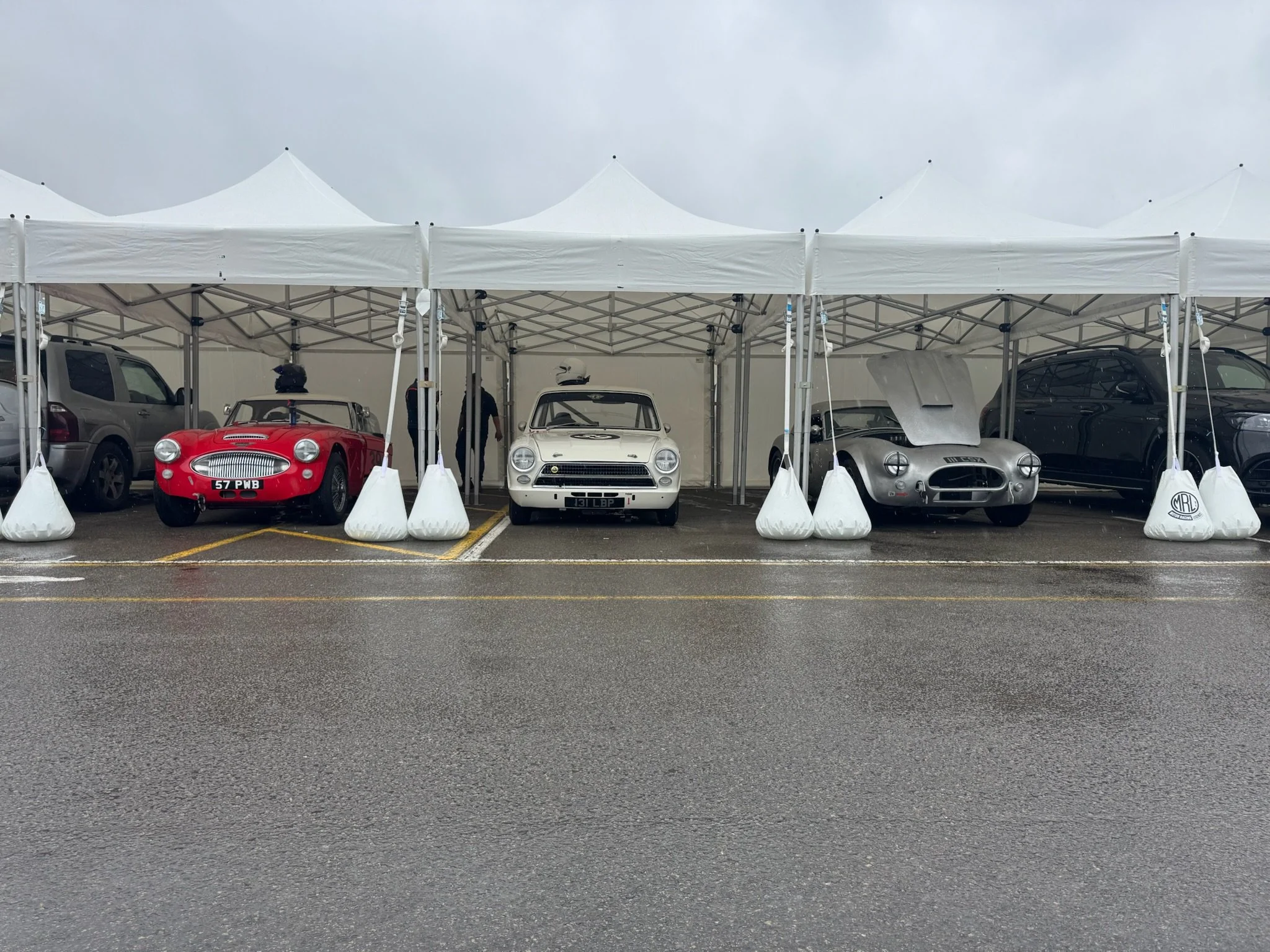 View of a row of race cars parked under a white canopy on a wet parking lot, with a gray cloudy sky overhead.