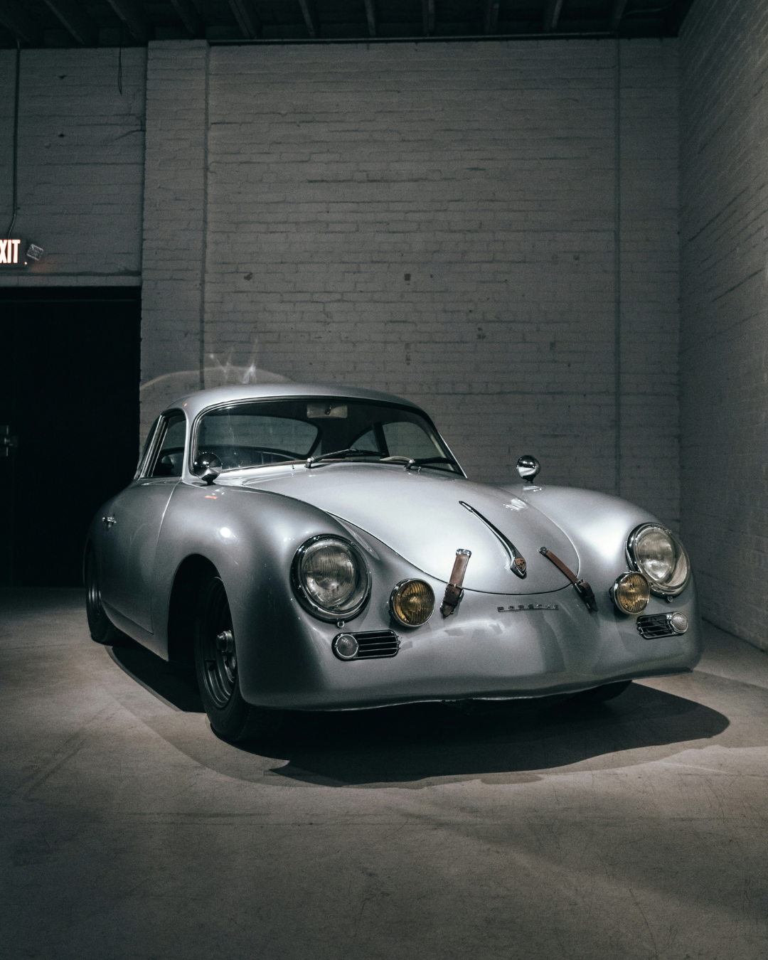 A vintage silver Porsche sports car on display in a dimly lit indoor setting with white brick walls.