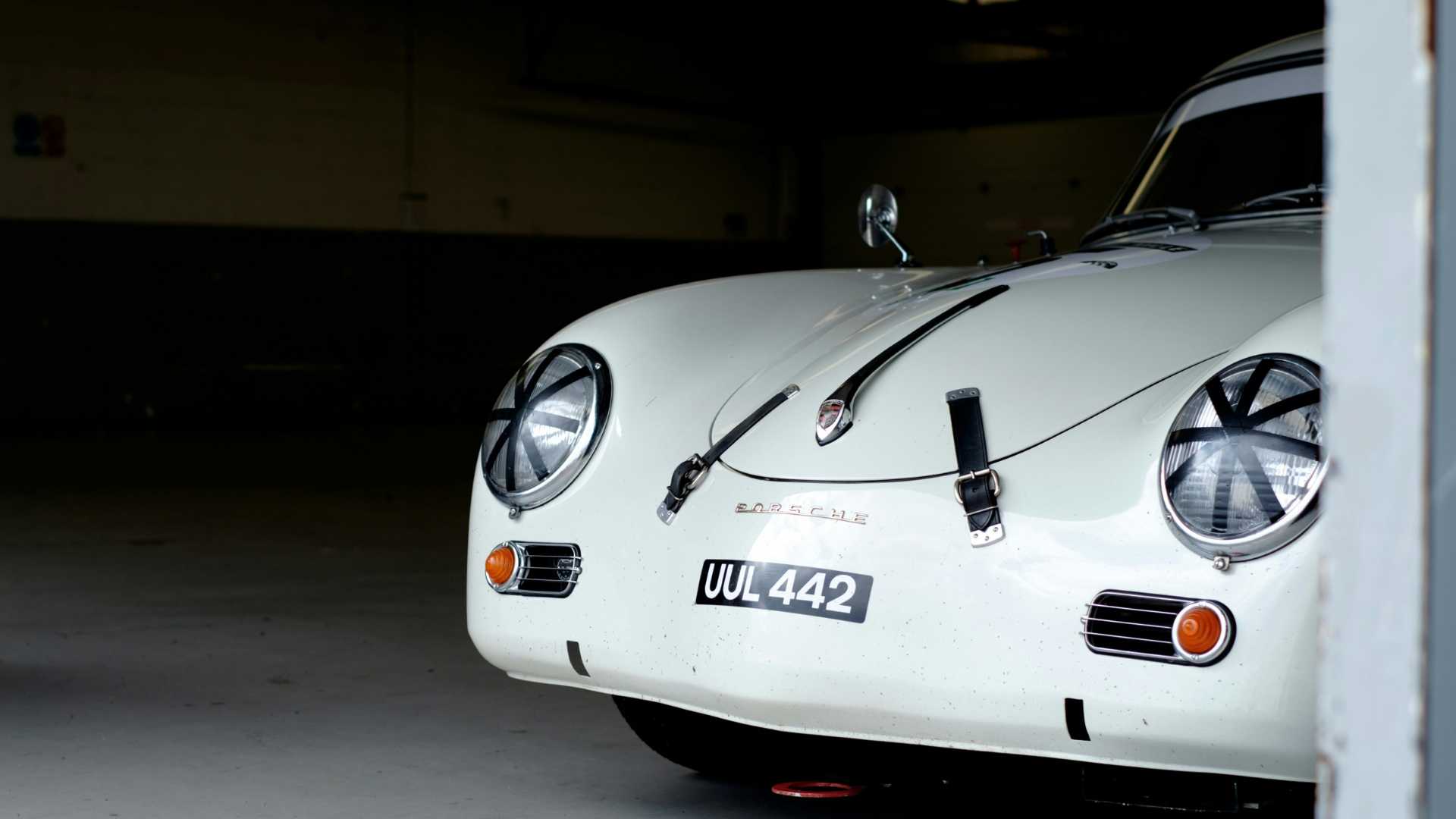 A classic white Porsche 356 sports car parked in a dimly lit garage, partially obscured by a wall on the right.