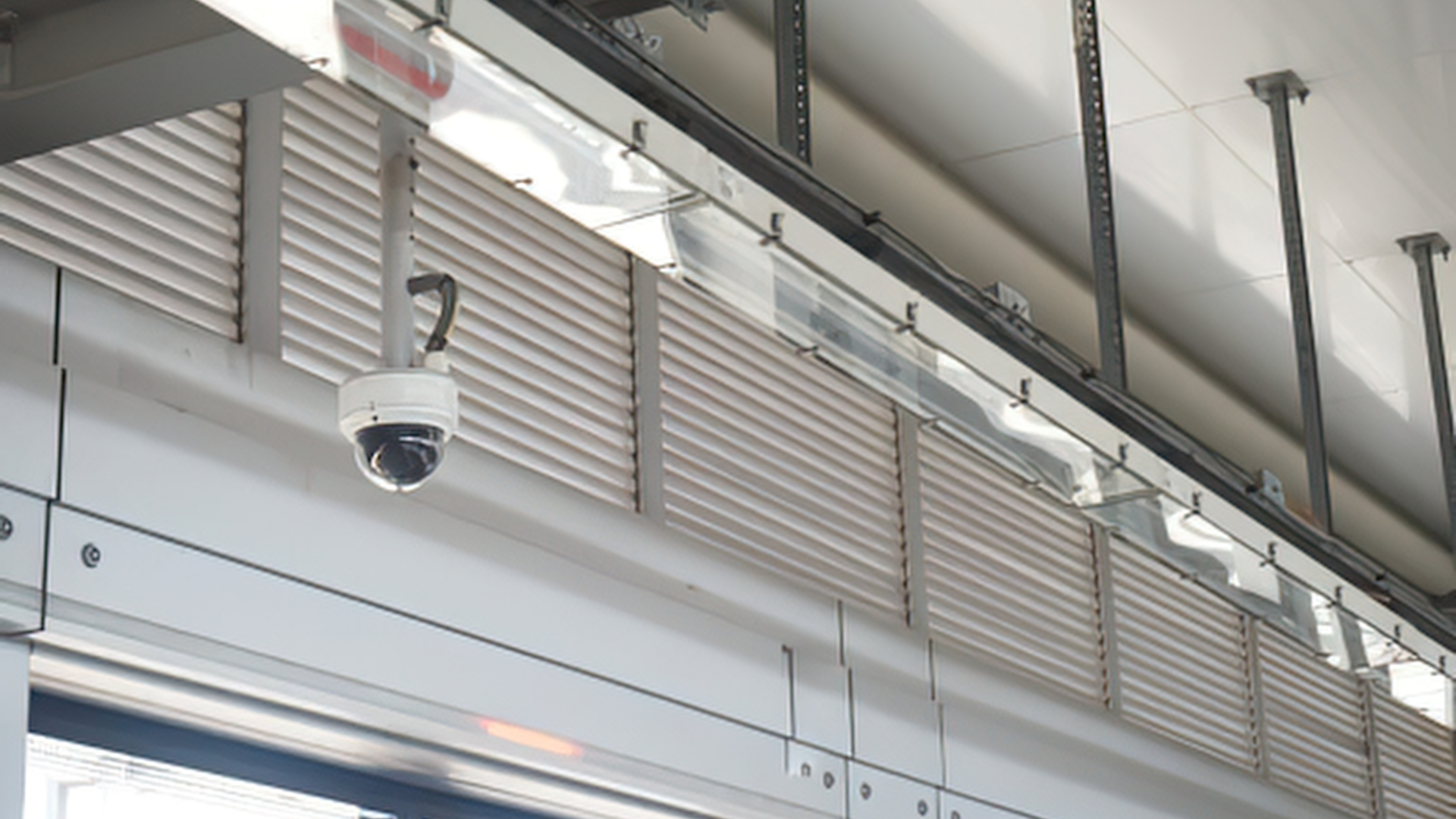 Ceiling with security camera, metal support beams, lighting fixtures, and horizontal ventilation panels in a modern building