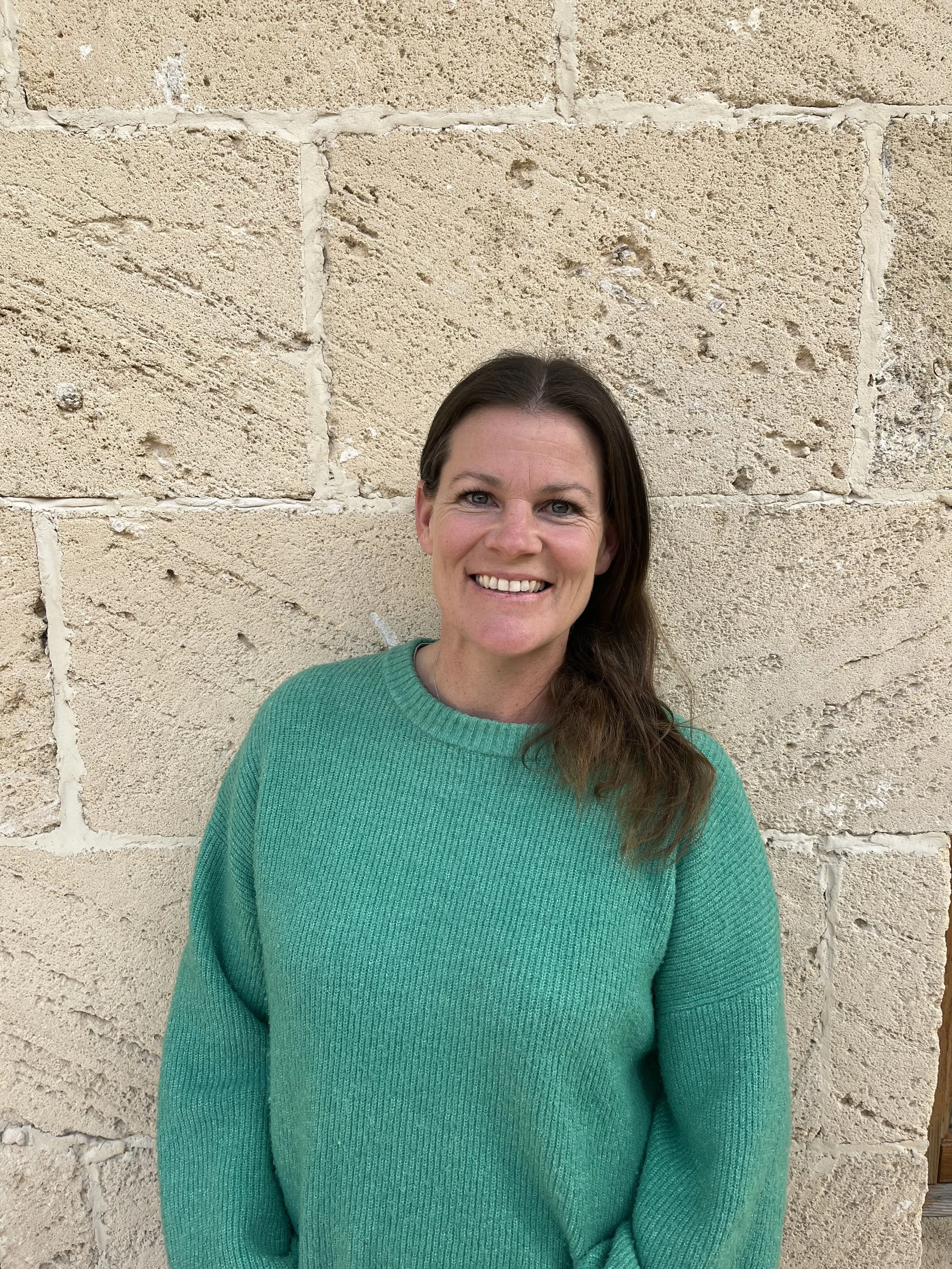 A woman with long brown hair, wearing a green sweater, smiling in front of a beige stone wall.