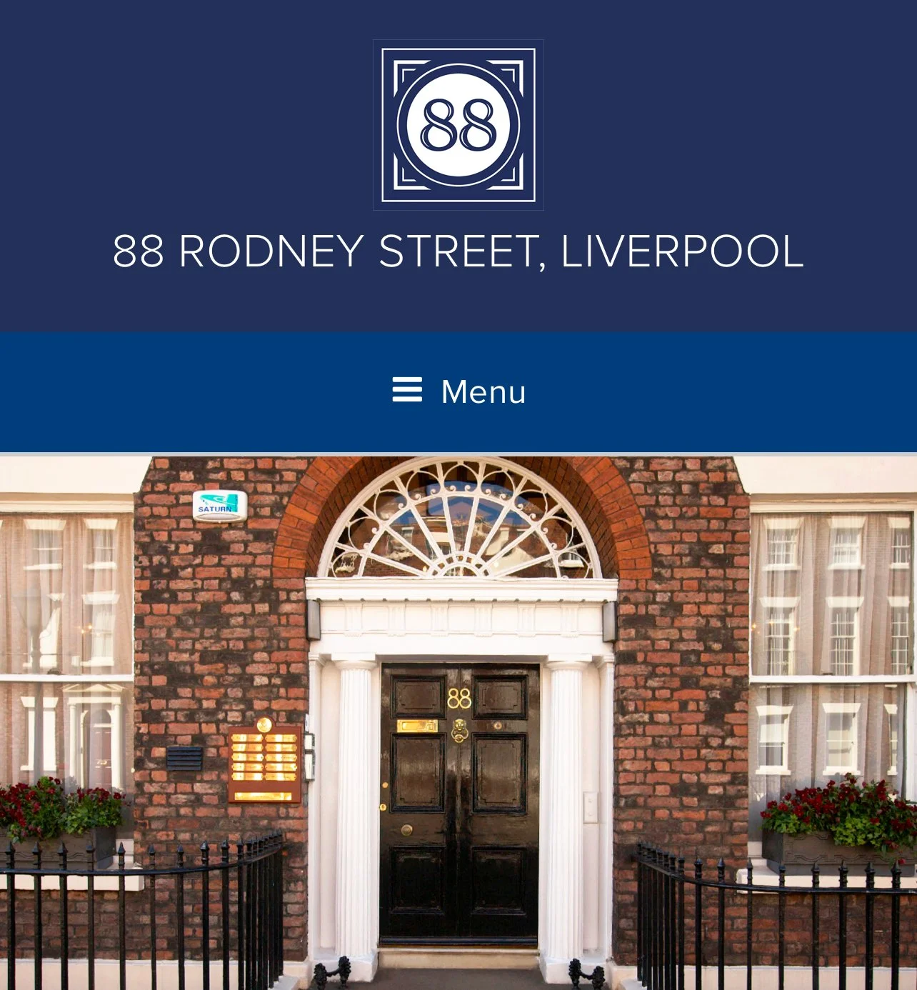 Street view of a brick building with a black door and white columns, marked with the number 88, with a window above the door and planters with flowers on either side.