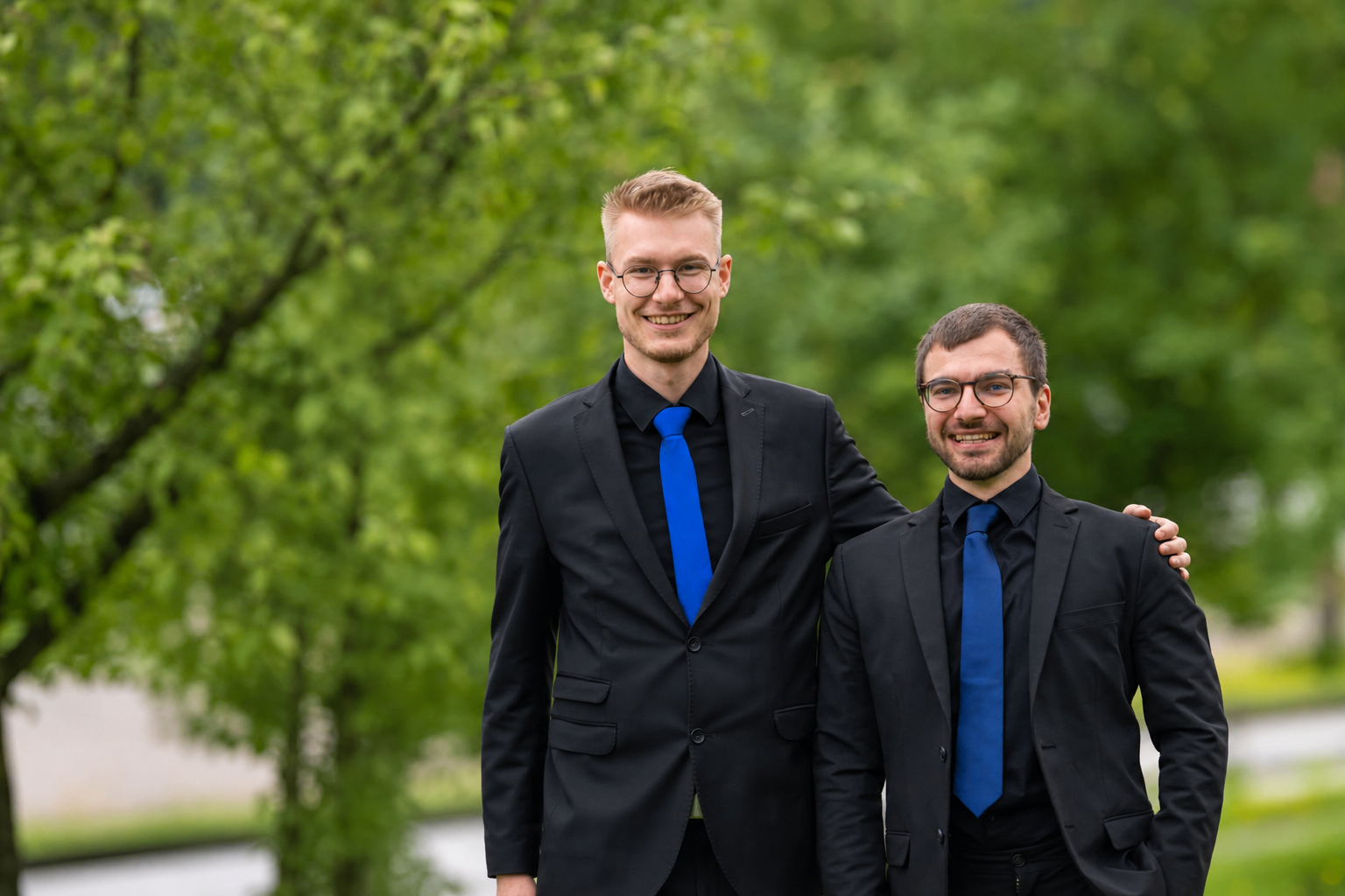 Zwei junge Männer in schwarzen Anzügen mit blauen Krawatten, die lächelnd im Freien vor grünen Bäumen stehen.