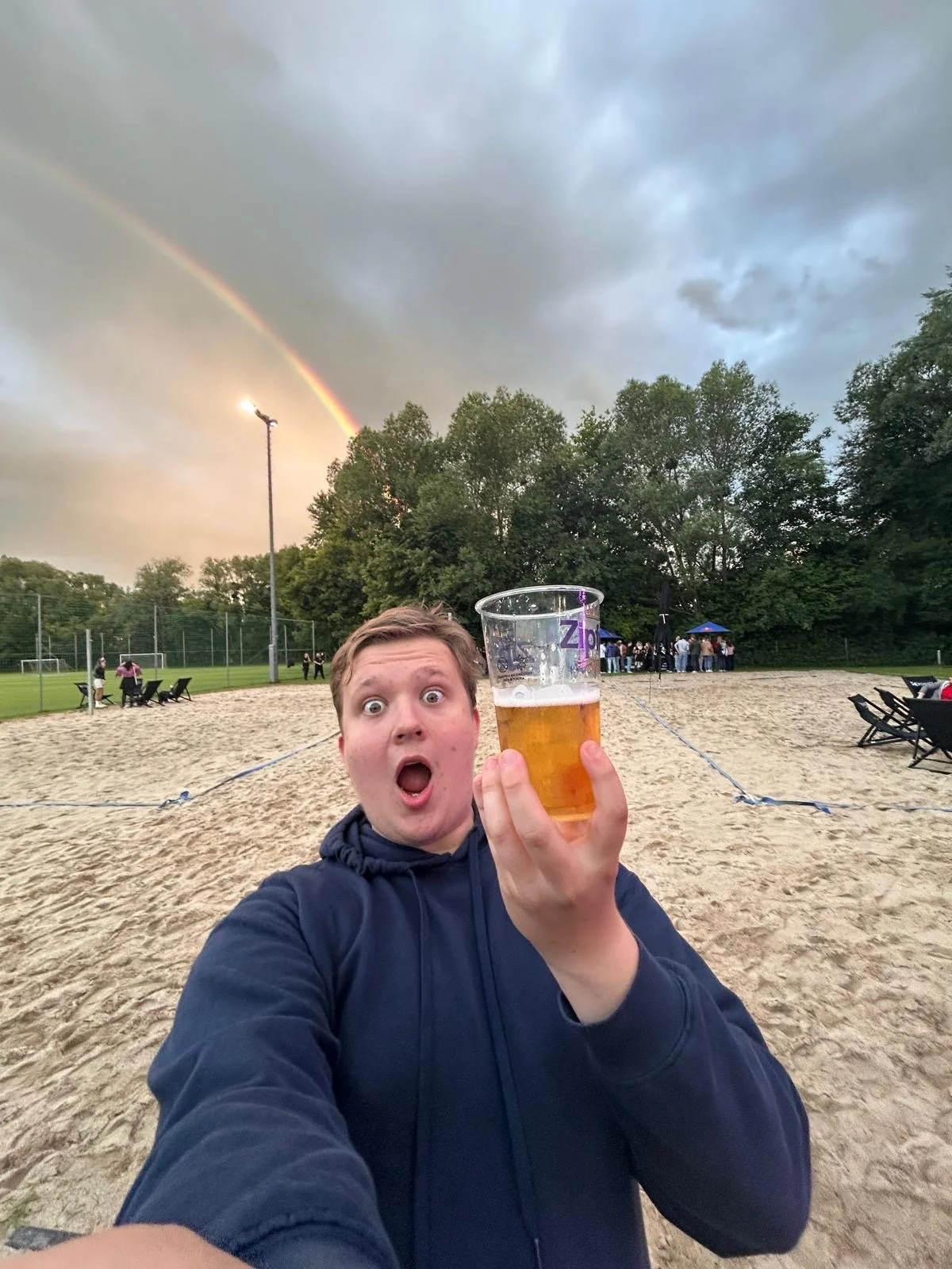 Ein junger Mann hält einen Becher Bier in der Hand, Blick überrascht, im Hintergrund eine Sandfläche, Menschen, Bäume, dunkler Himmel mit Regenbogen und Wolken.