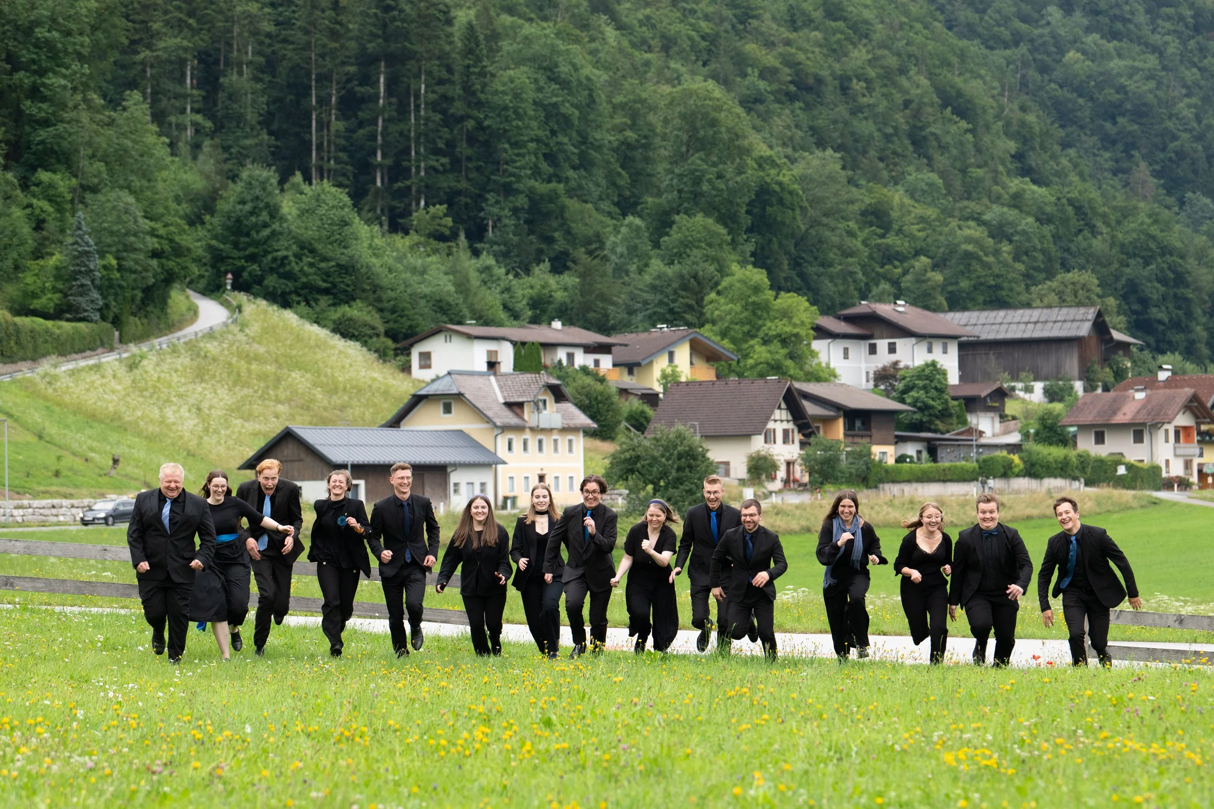 Eine Gruppe fröhlicher Menschen in formeller Kleidung läuft über eine grüne Wiese vor einem Dorf mit Häusern, umgeben von Bäumen.