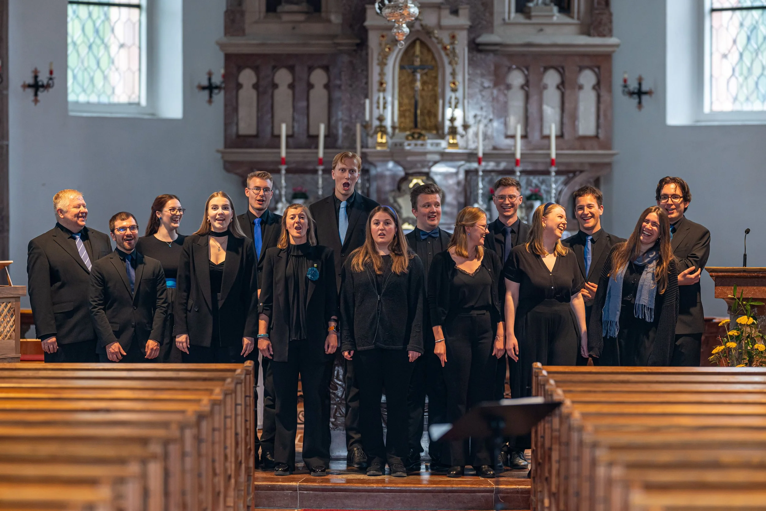 Chor von jungen Männern und Frauen, die in einer Kirche singen, getragen in formellen schwarzen Anzügen und Kleidern, mit einem geschnitzten Altar im Hintergrund und Ausschmückungen.