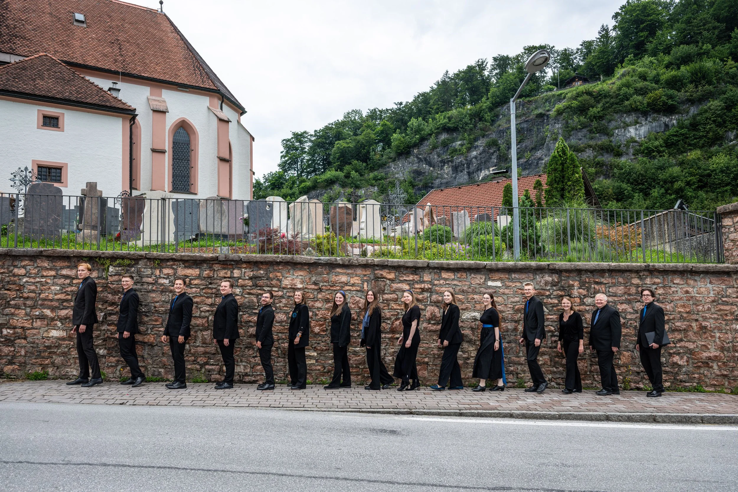 Eine Gruppe von Menschen in schwarzen Anzügen läuft in einer Reihe entlang einer Mauer. Im Hintergrund befindet sich ein Friedhof mit Grabsteinen, einem weißen Gebäude mit roten Dachschindeln und einer grünen, bewaldeten Hügel.