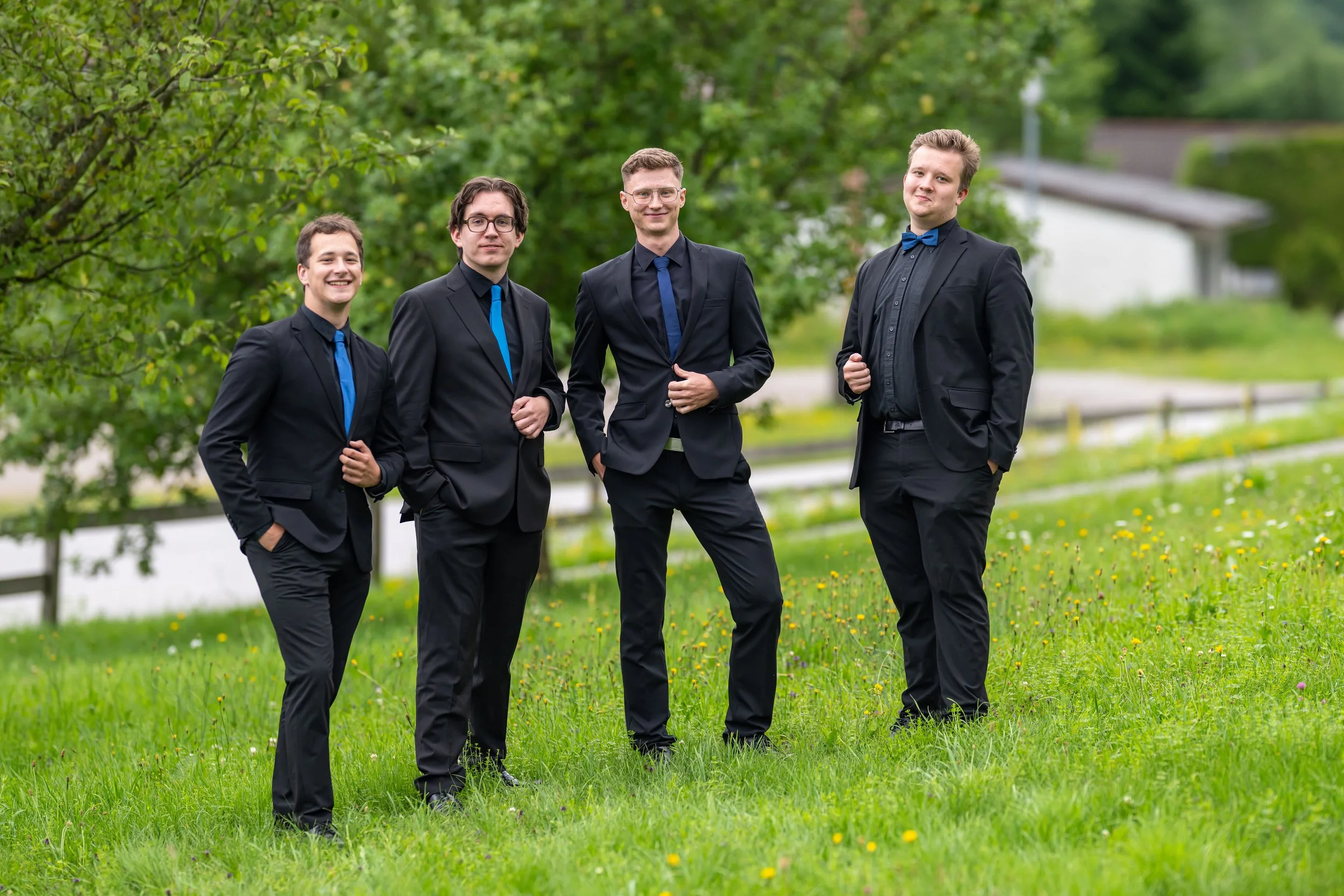 Vier junge Männer in schwarzen Anzügen mit blauen Accessoires stehen auf einer grünen Wiese vor Bäumen und einem Wasserweg.