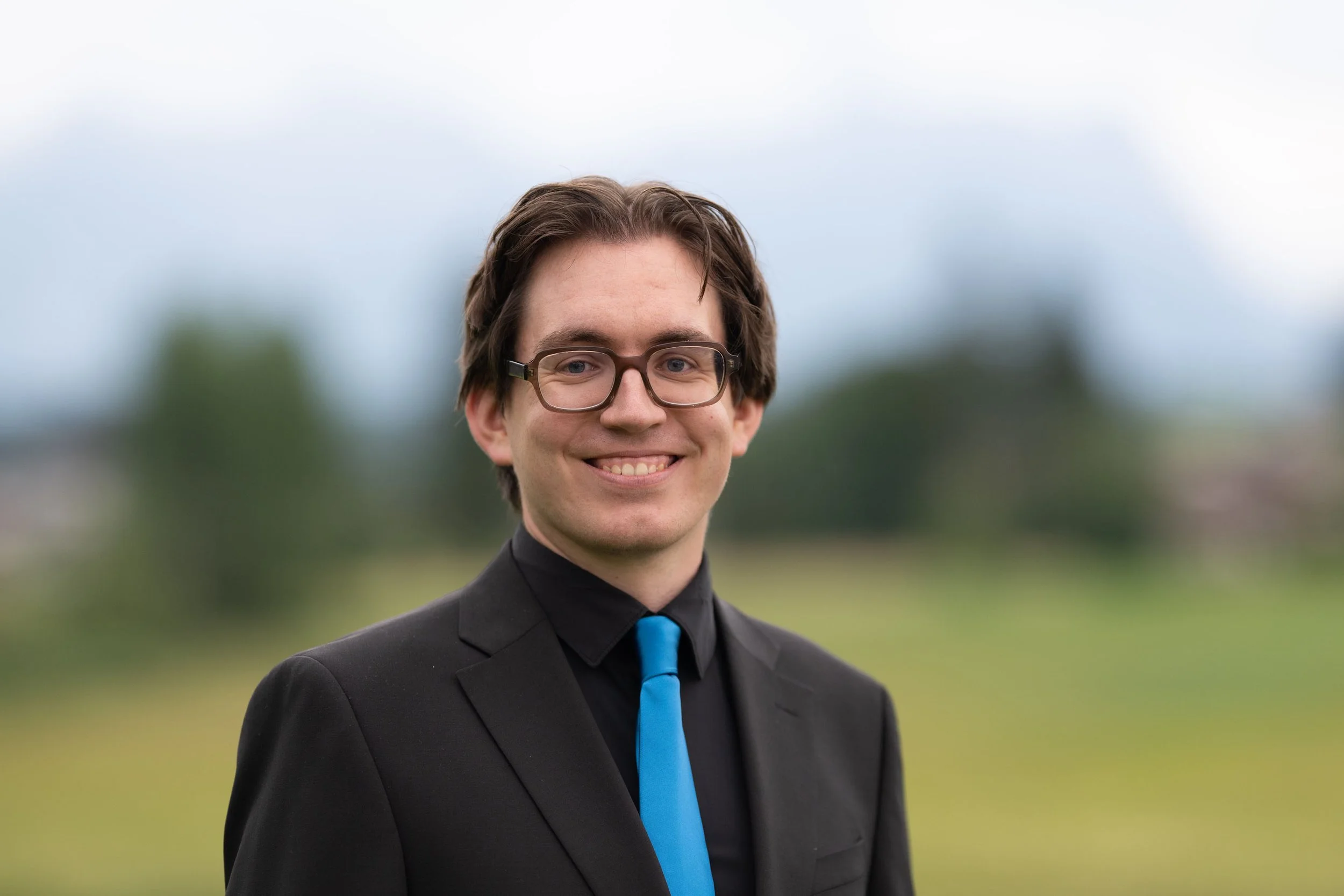 Junge Mann trägt schwarze Kleidung, blaue Krawatte und Brille, steht im Freien mit unscharfem grünen Hintergrund, lächelt in die Kamera.
