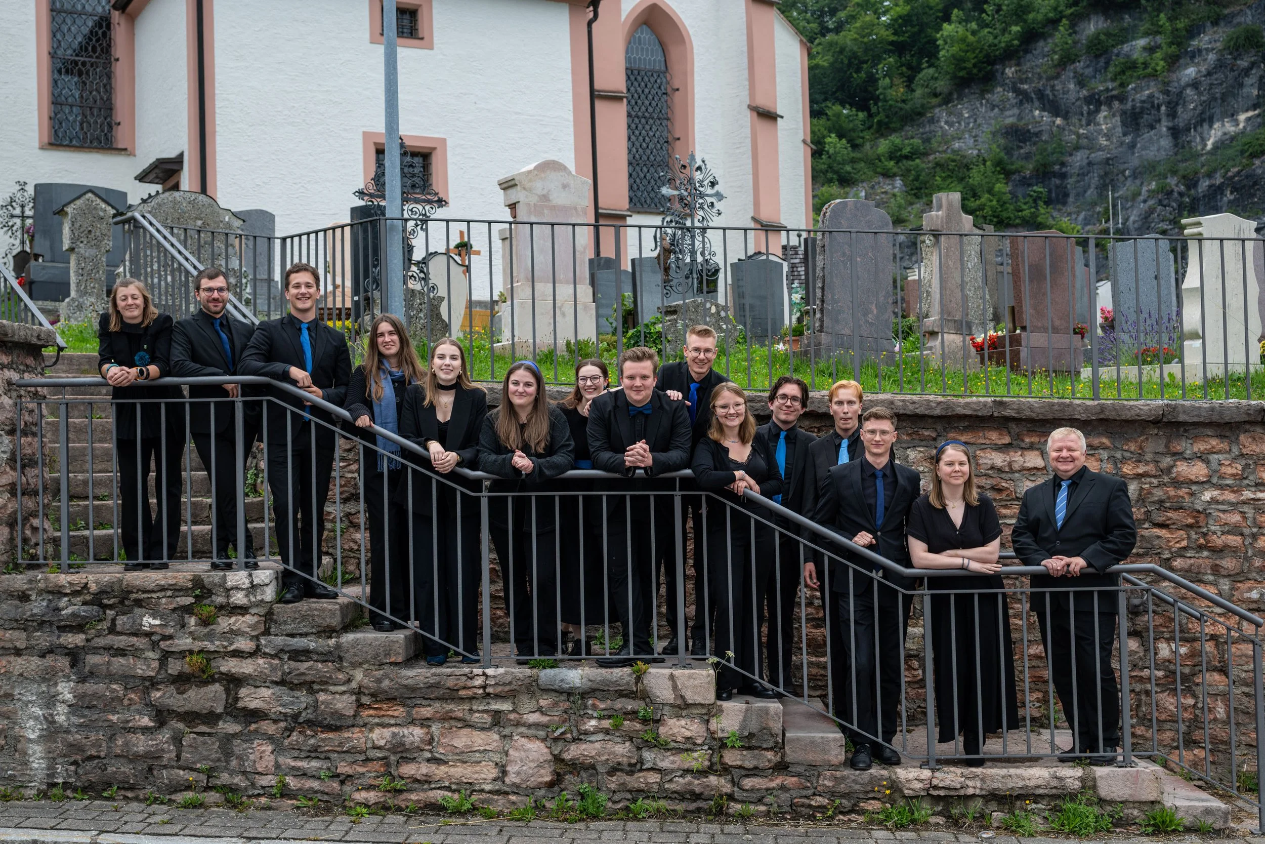 Eine Gruppe junger Menschen in formeller Kleidung steht vor einer alten Steinmauer, eine kleine Anzahl sitzt, einige stehen, vor einer Kirche mit einem Friedhof im Hintergrund.