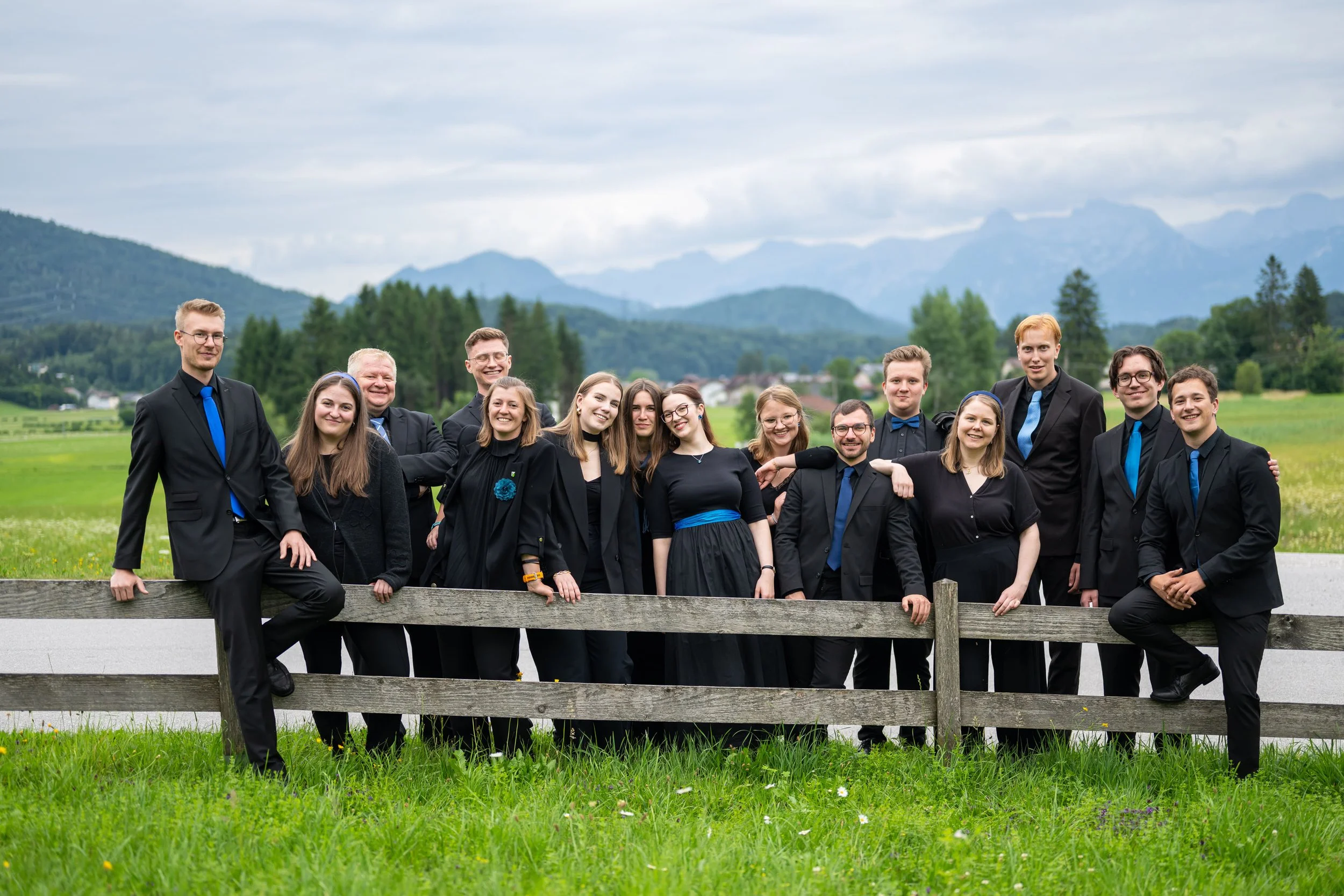 Eine Gruppe junger Menschen in formeller Kleidung steht außerhalb auf einer Wiese in einer ländlichen Gegend mit Bergen im Hintergrund. Sie sind lächelnd und posieren für die Kamera.