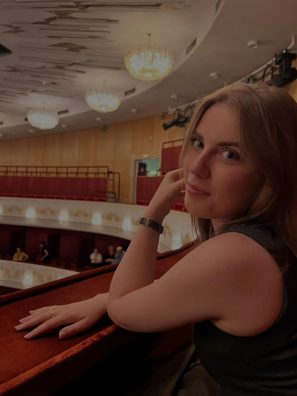 Eine junge Frau mit braunen Haaren sitzt in einem Theater oder Opernhaus, schaut in die Kamera und lächelt leicht. Die Kulisse zeigt eine elegante Innenarchitektur mit Kronleuchtern und einer Balustrade im Hintergrund.