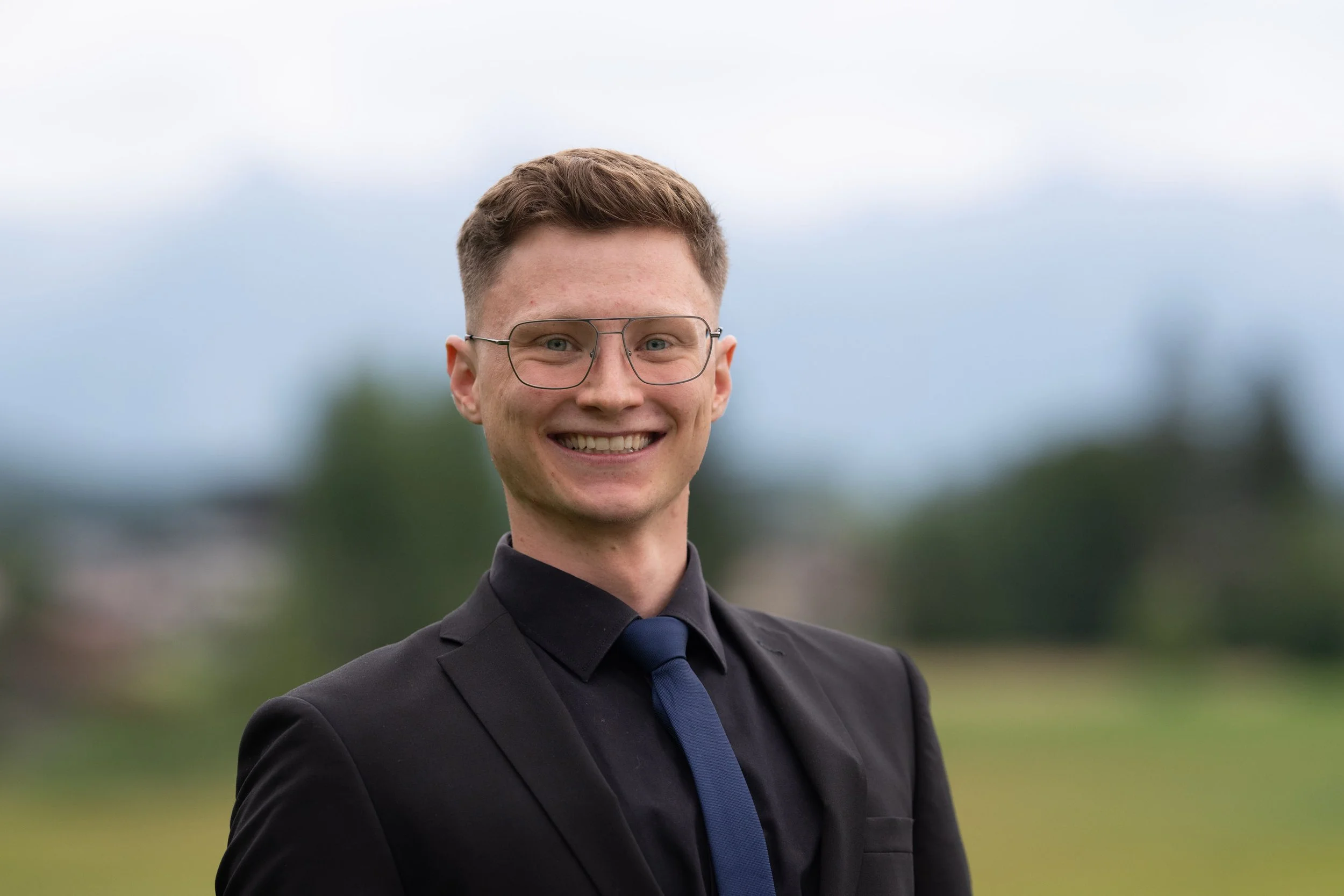 Ein junger Mann trägt eine schwarze Anzugsjacke, eine schwarze Krawatte und eine dunkle Krawatte. Er trägt eine große Brille und hat braunes, kurz geschnittenes Haar. Der Hintergrund zeigt eine unscharfe, grüne Landschaft und einen meist bewölkten Himmel.