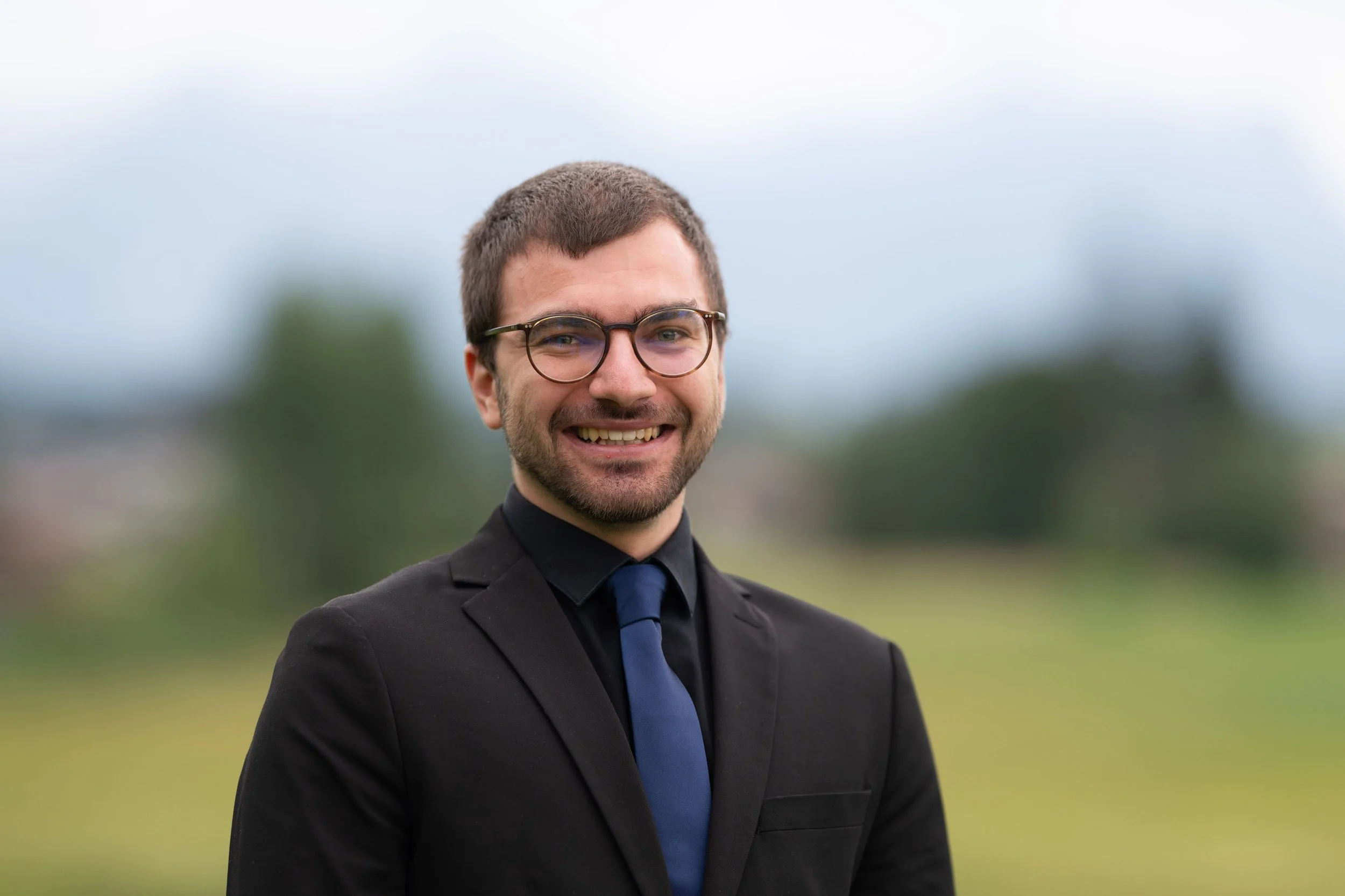 Ein junger Mann in einem schwarzen Anzug mit blauer Krawatte und Brille, lächelnd im Freien, mit einer bewölkten Himmel im Hintergrund.