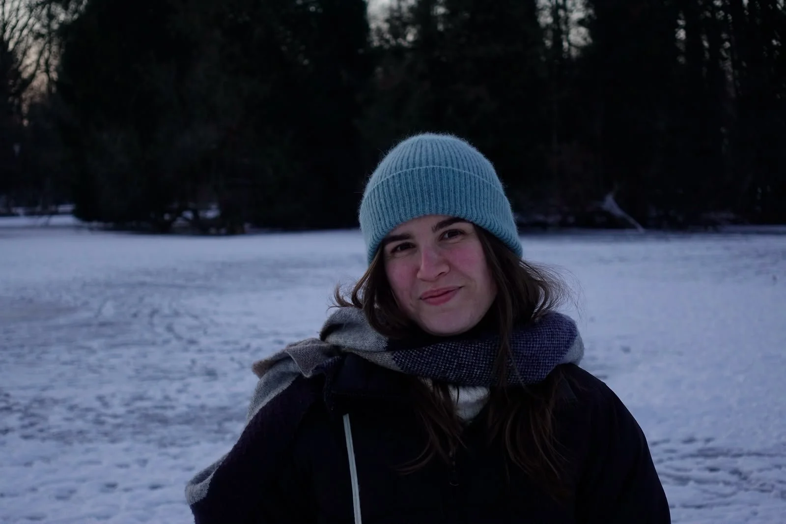 Junge Frau mit blauer Mütze und dunklem Wintermantel im Schnee, im Hintergrund Tannenbäume bei Dämmerung.