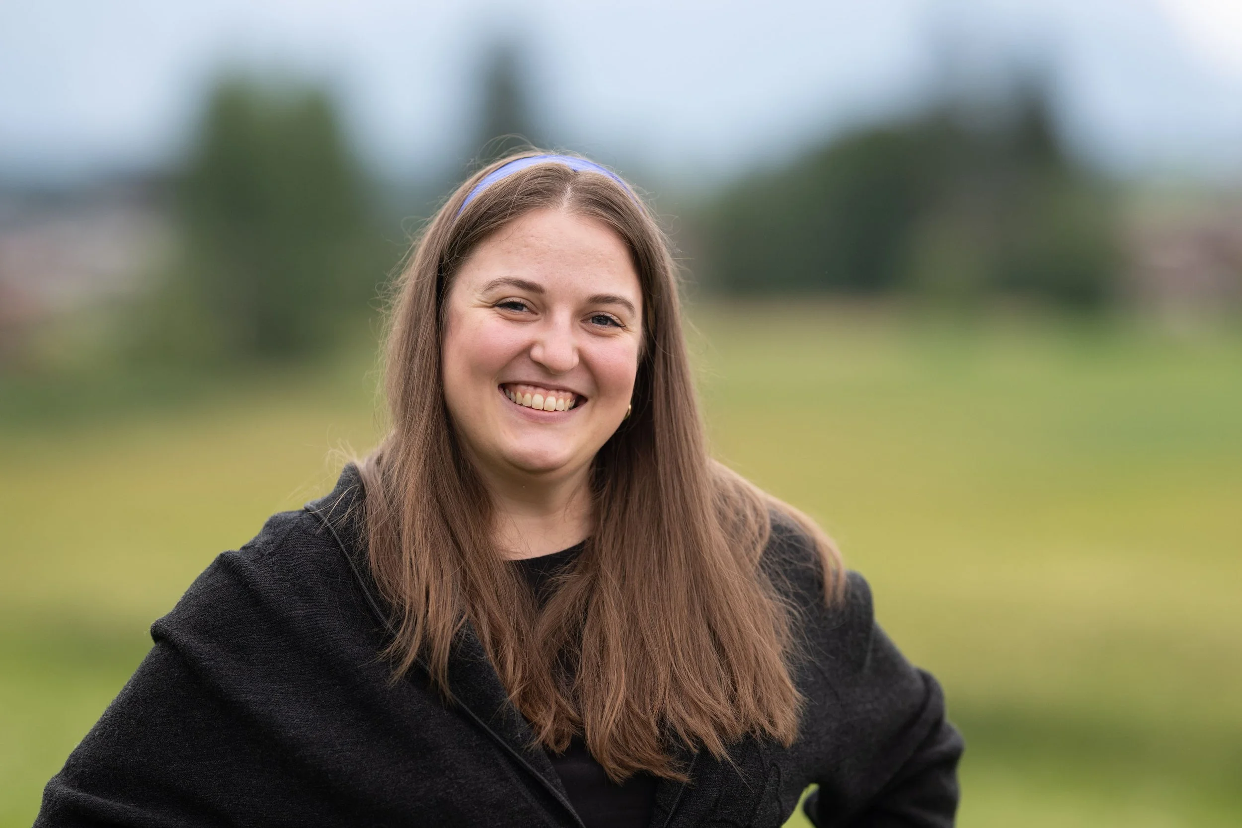 Ein lächelndes junges Mädchen mit langen braunen Haaren, Haare mit blauer Stirnband, trägt schwarze Jacke, draußen in einer grünen, leicht unscharfen Umgebung.