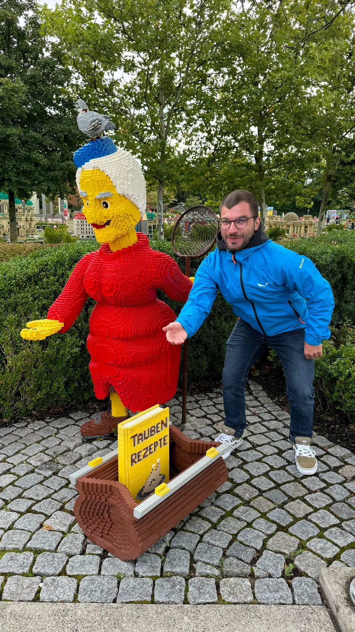Ein Mann in blauer Jacke posiert neben einer Figur, die aus LEGO-Steinen gebaut ist. Die LEGO-Figur stellt eine Frau mit rotem Kleid, gelbem Haar und einem weißen Haarschopf dar. Es gibt auch einen LEGO-Block mit dem Text 'TAUBEN REZEPTE'.