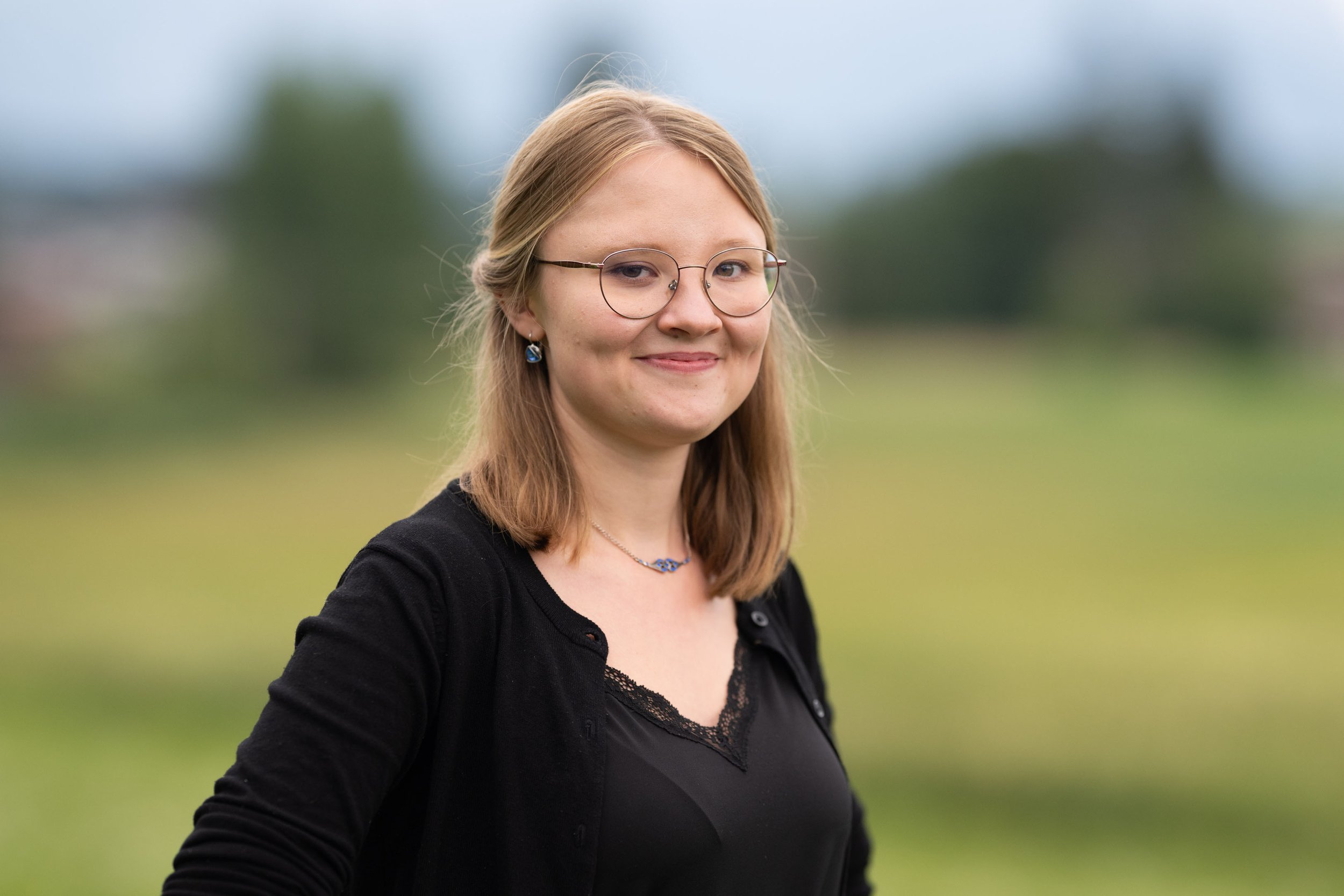 Junge Frau mit Brille, braunen Haaren, dunklem Oberteil und lila Ohrringen lächelt im Freien, im Hintergrund ist eine grüne Wiese und der Himmel zu sehen.