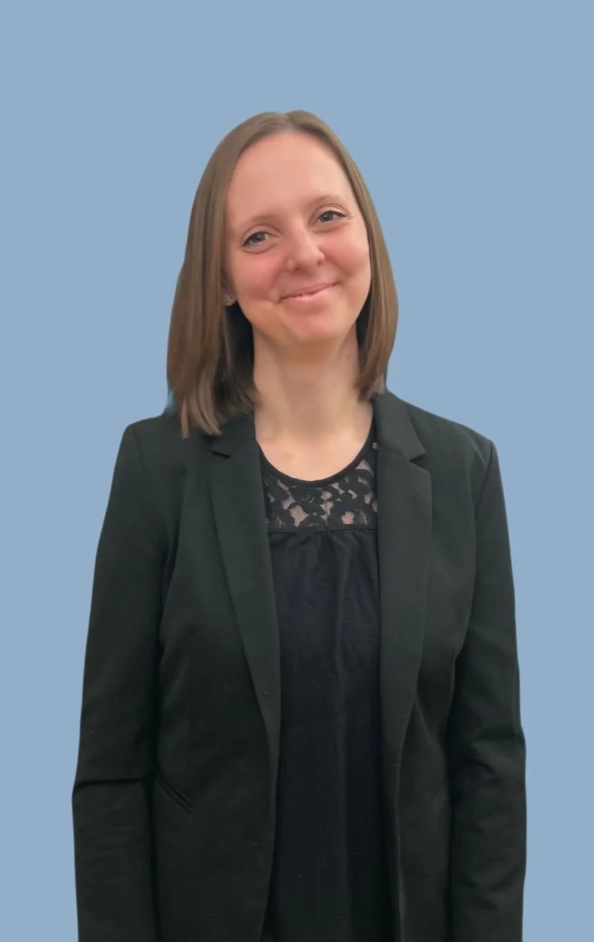 Junge Frau mit braunen Haaren, schwarzem Blazer und schwarzem Top, lächelnd vor blauem Hintergrund.