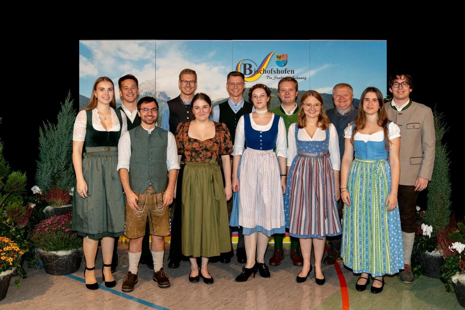 Gruppe von Menschen in traditioneller bayerischer Tracht vor einem Hintergrund mit Berglandschaft und Logo der Stadt Bischofshofen.