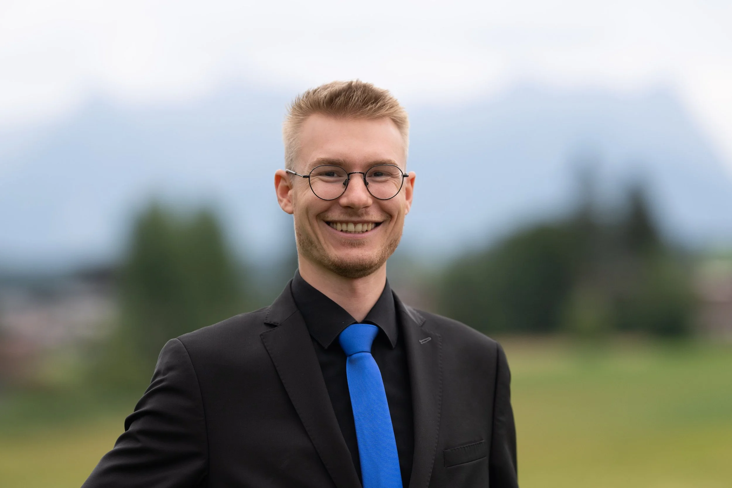 Ein junger Mann trägt einen schwarzen Anzug mit blauer Krawatte und hat kurze, blonde Haare, er lächelt vor einem bewölkten Himmel im Freien.