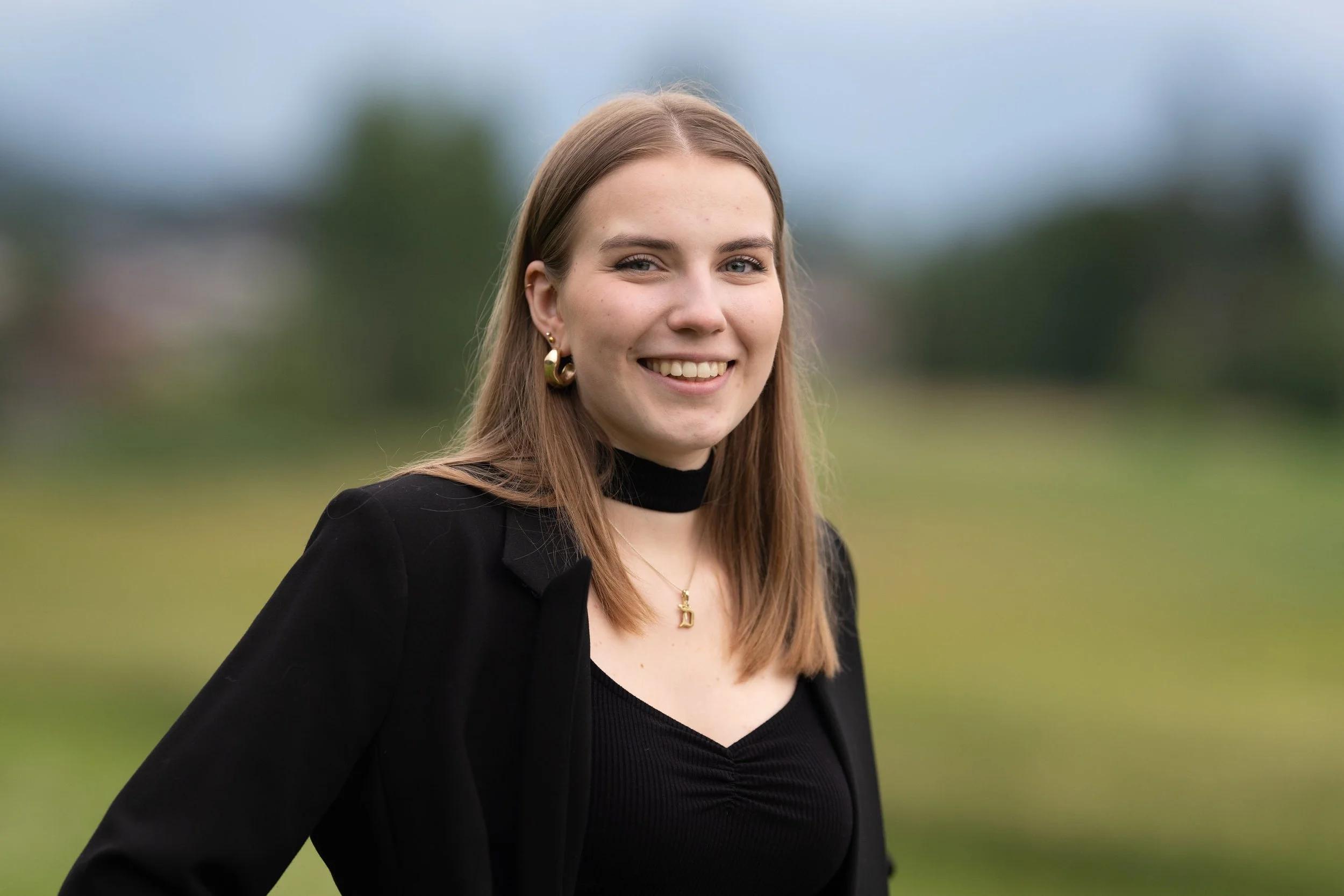 Lächelnde junge Frau mit langen braunen Haaren, goldene Ohrringe und Halskette, trägt schwarze Jacke und Top, steht im Freien mit grünem Hintergrund.