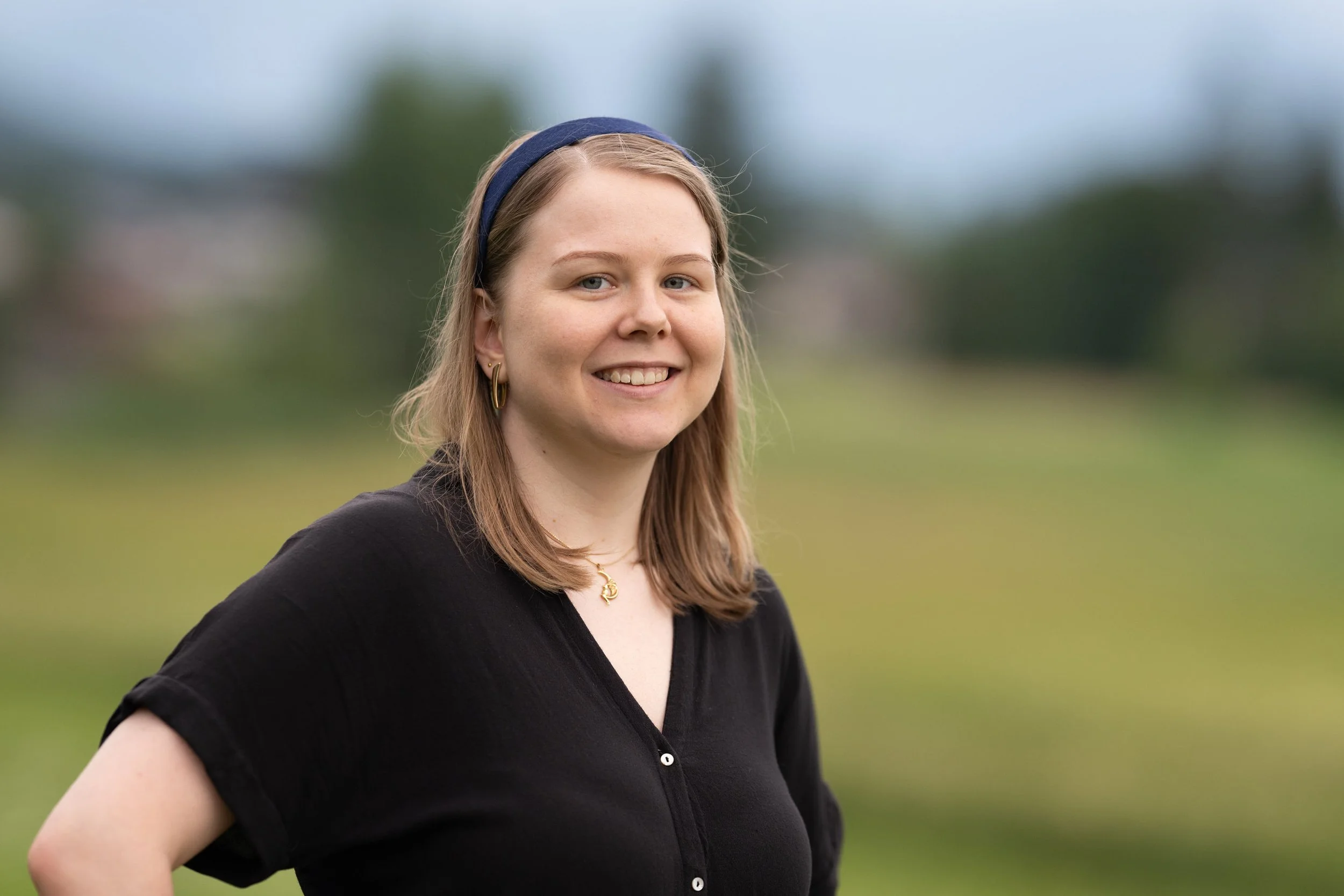 Eine junge Frau mit braunen Haaren, Ohrringen und einem blauen Haarband, die draußen im Grünen lächelt.