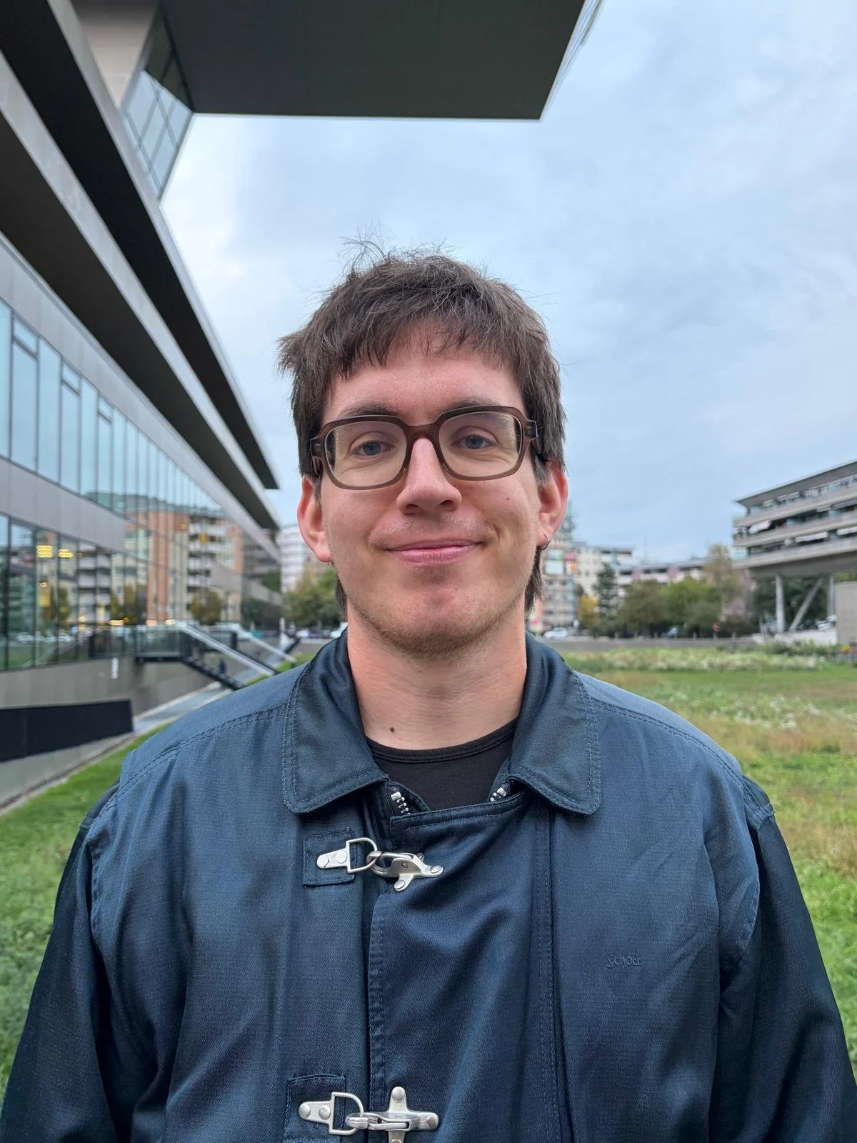 Ein junger Mann mit Brille, dunkler Jacke und Lächeln steht im Freien vor modernen Gebäuden und Grünflächen.