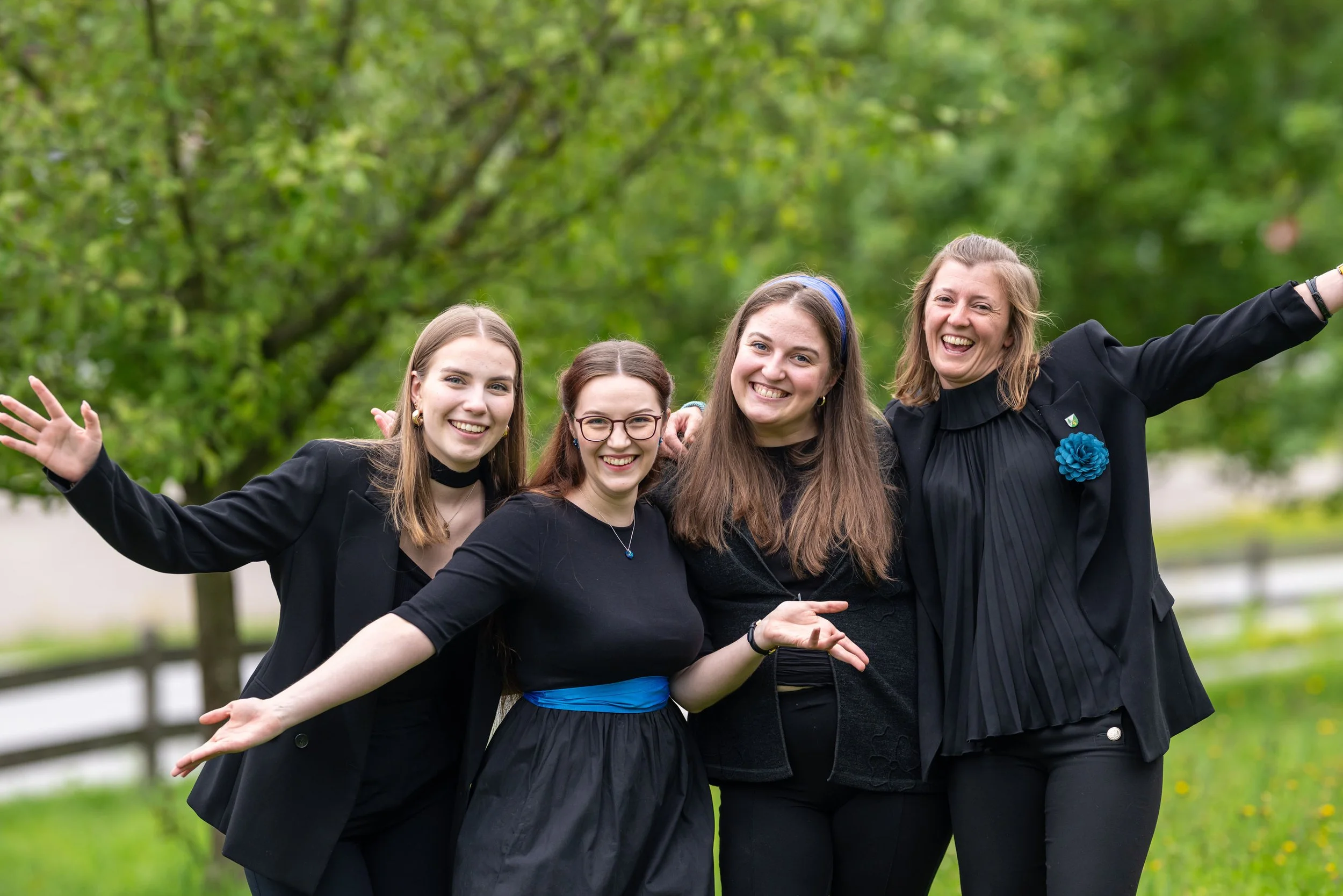 Fünf lachende Frauen in schwarzen Outfits, die im Freien vor grünen Bäumen Spaß haben.