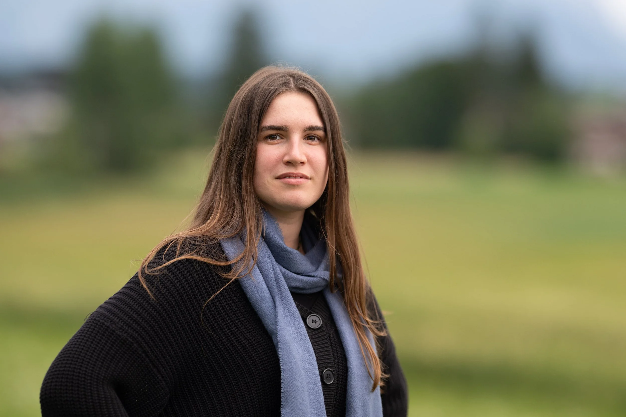 Junge Frau mit braunen Haaren, grauem Schal und schwarzer Jacke steht im Freien vor grüner Landschaft mit Bäumen, bewölkter Himmel im Hintergrund.