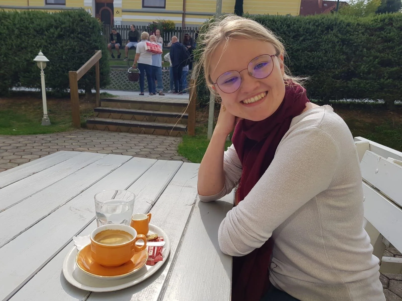 Eine lachende Frau mit Brille sitzt an einem weißen Tisch im Freien, trinkt Kaffee und hat einen weiteren Kaffee auf einem Tablett vor sich.
