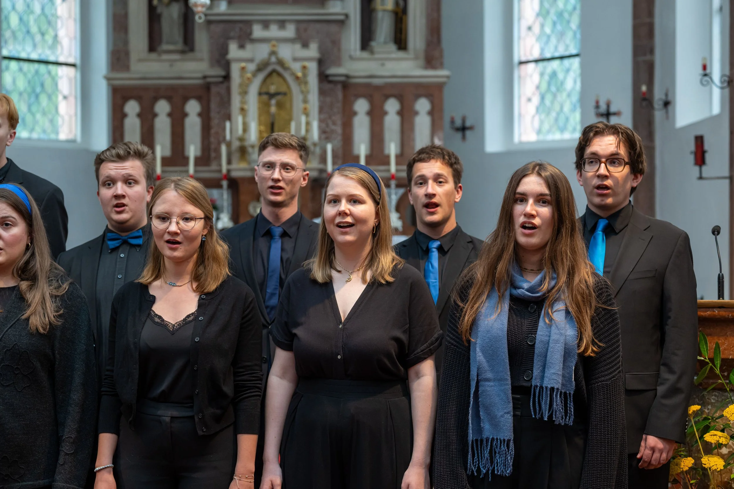Jugendlicher Chor singt in einer Kirche, mit Altar im Hintergrund und Fenster mit Gittern. Alle tragen schwarze Kleidung mit blauen Accessoires.