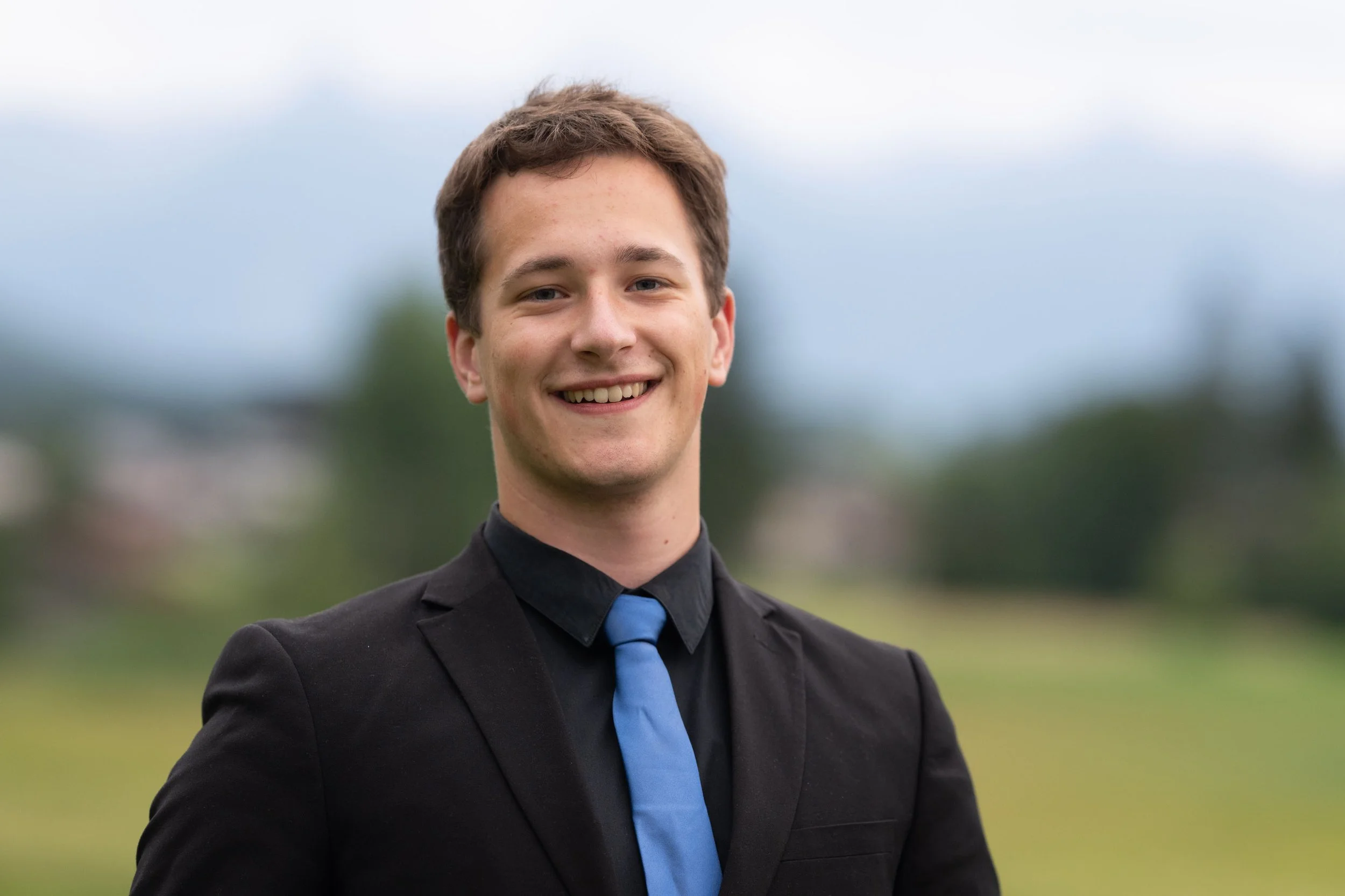 Junger Mann in formeller schwarzer Anzug mit blauer Krawatte, lächelnd, im Freien auf einer Wiese bei bewölktem Himmel.