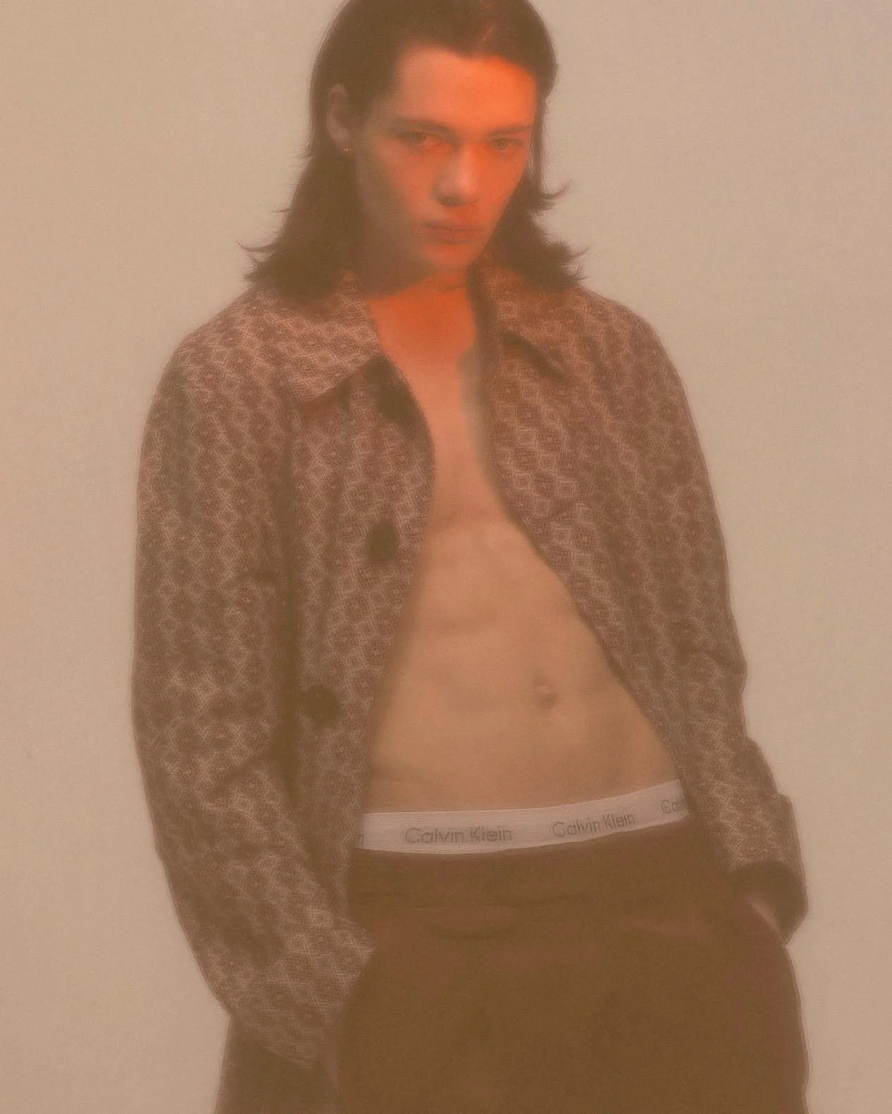 A young man with long dark hair, partially unbuttoned patterned shirt, black Calvin Klein underwear, and dark pants, standing against a plain background with a serious expression.