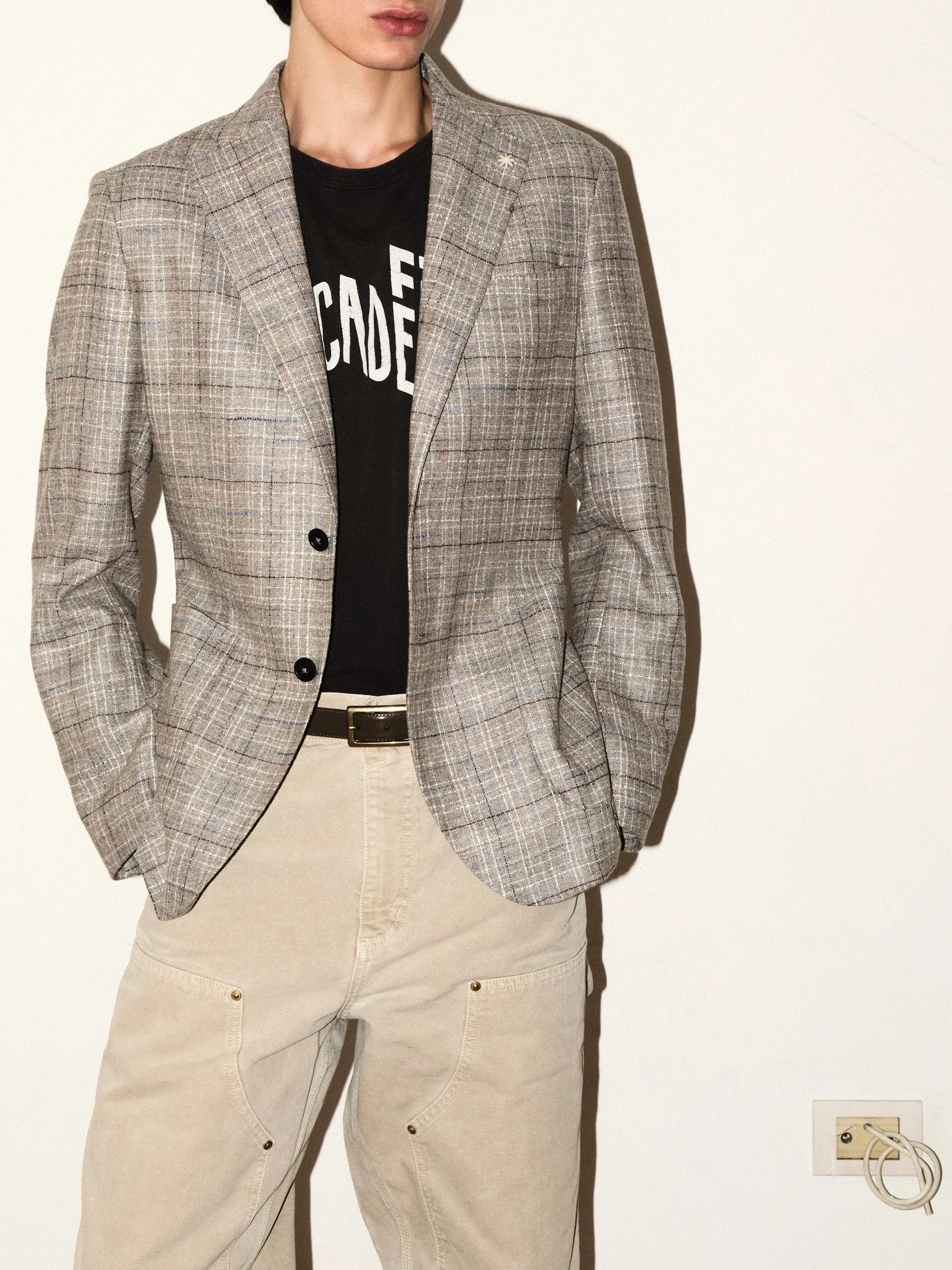 Person wearing a plaid blazer over a black graphic t-shirt and beige pants, standing against a plain wall, with a power outlet and cords on the right side of the image.