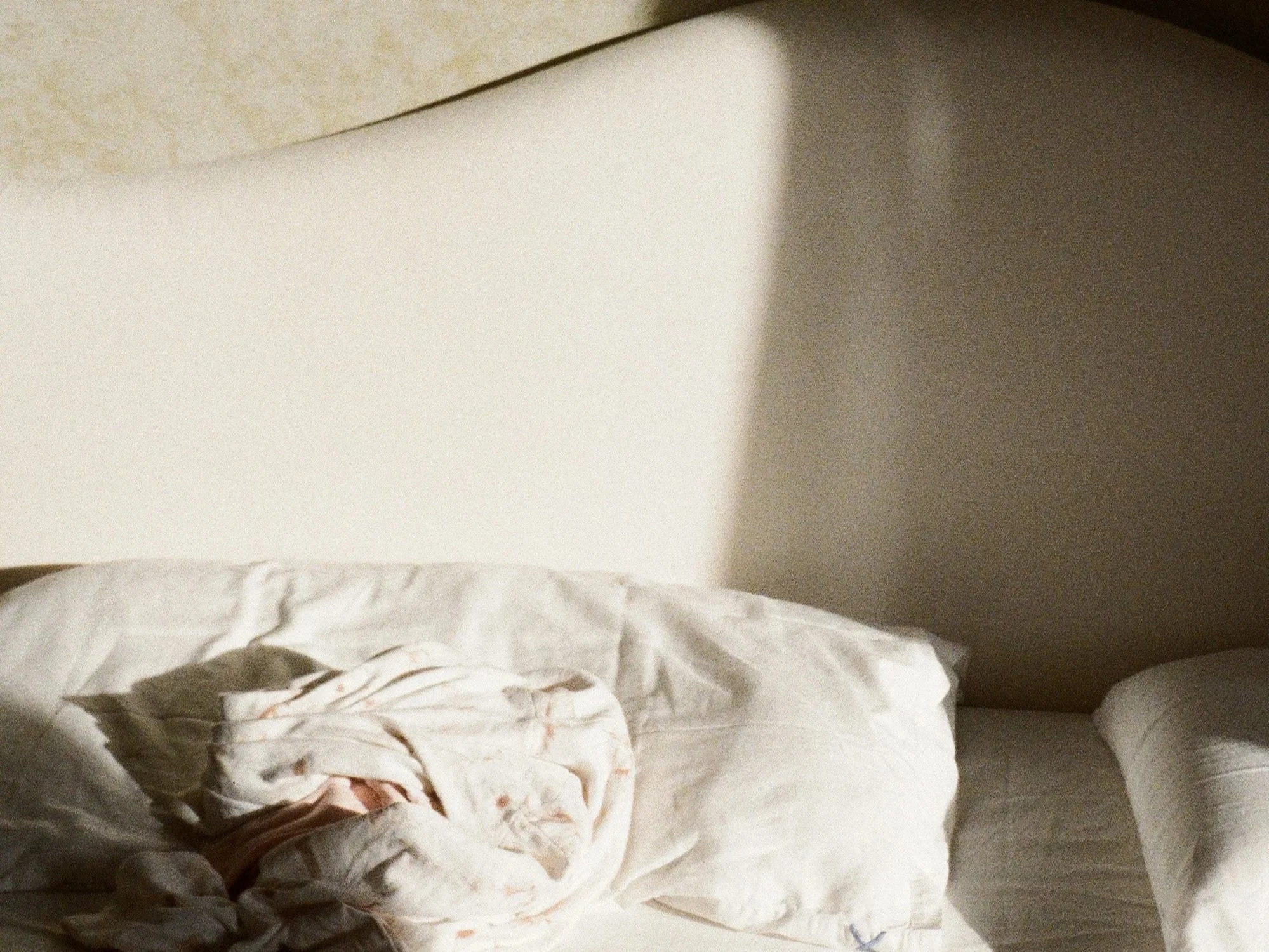 A white bed with a crumpled, patterned blanket at the foot of the bed, against a neutral-colored wall with a visible shadow.