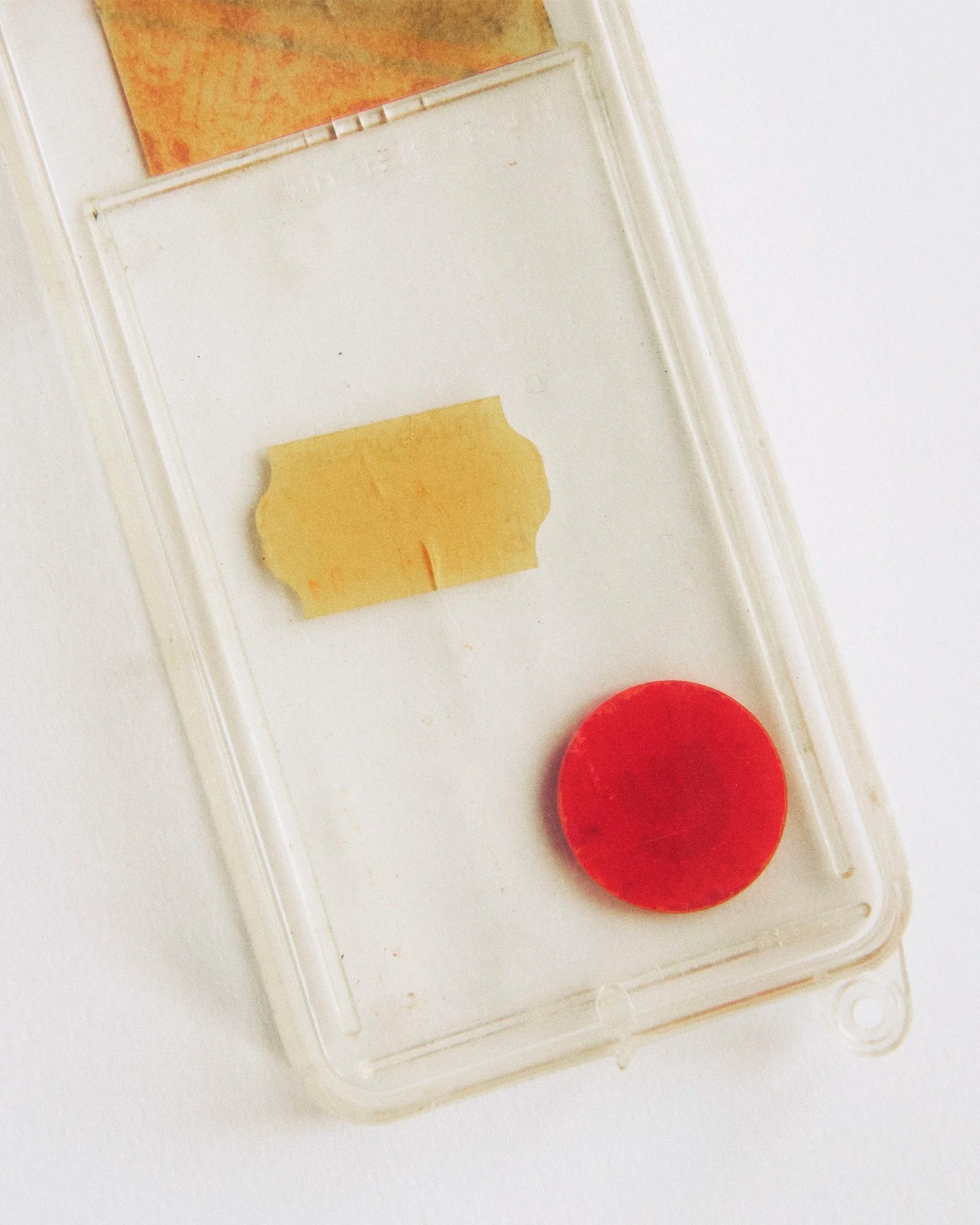 Close-up of a white plastic container holding a yellow waxy substance in a small piece, a red round object, and a partially visible orange object at the top.