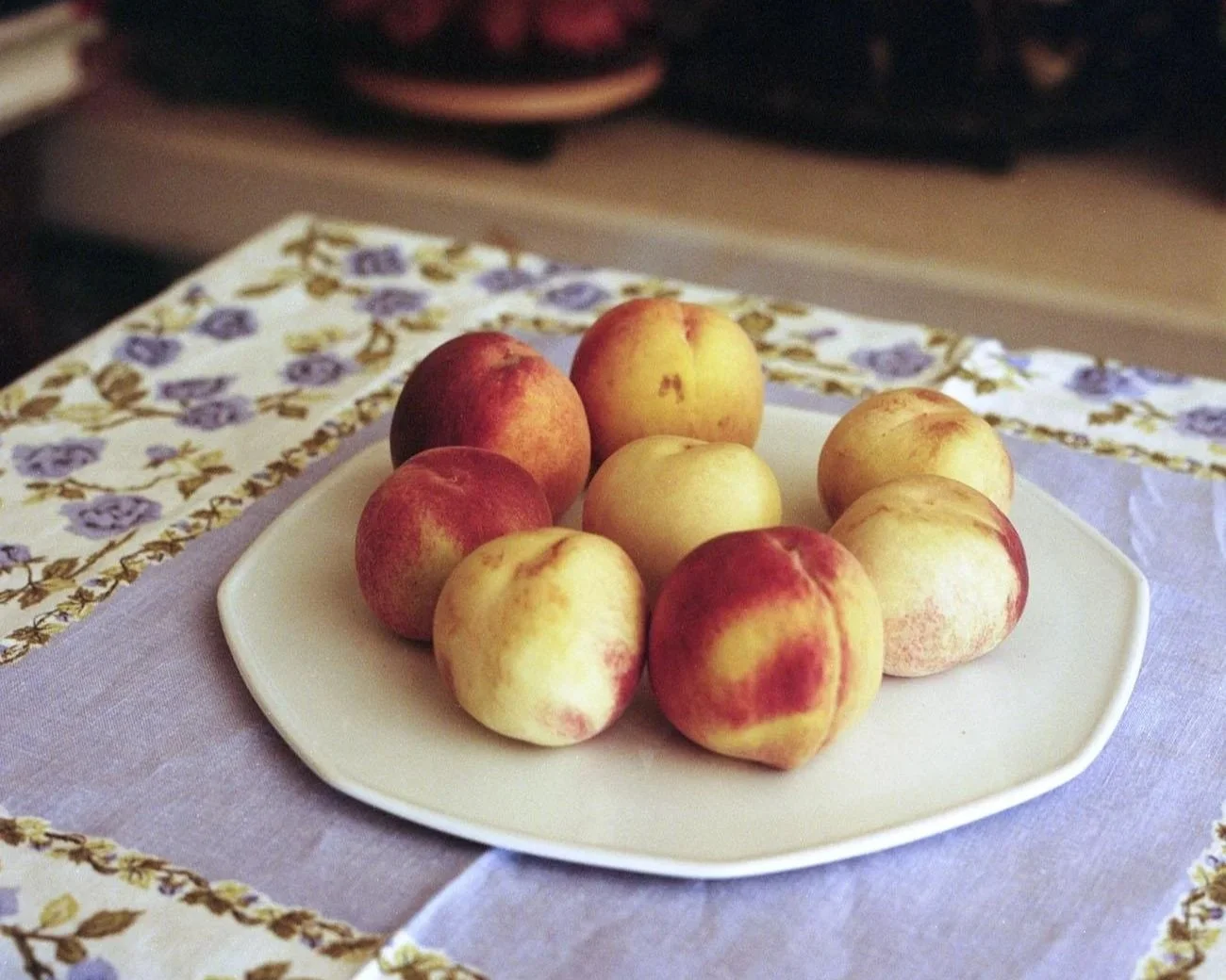 A white plate with ten peaches resting on a table decorated with a floral tablecloth featuring purple flowers and green leaves.