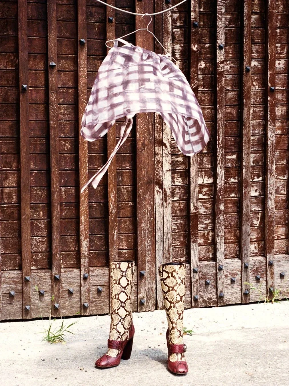 An outdoor scene with a wooden fence. Instead of a person, there are legs wearing snake-print stockings and red high-heeled shoes, with a plaid shirt draped and hanging from a hanger above.