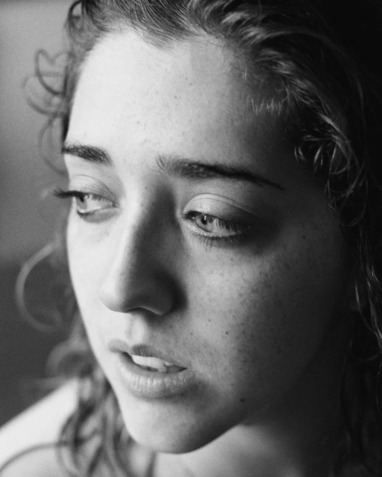 A close-up black and white photo of a woman's face, with her eyes looking to the side; her skin is smooth, and her hair has loose curls.