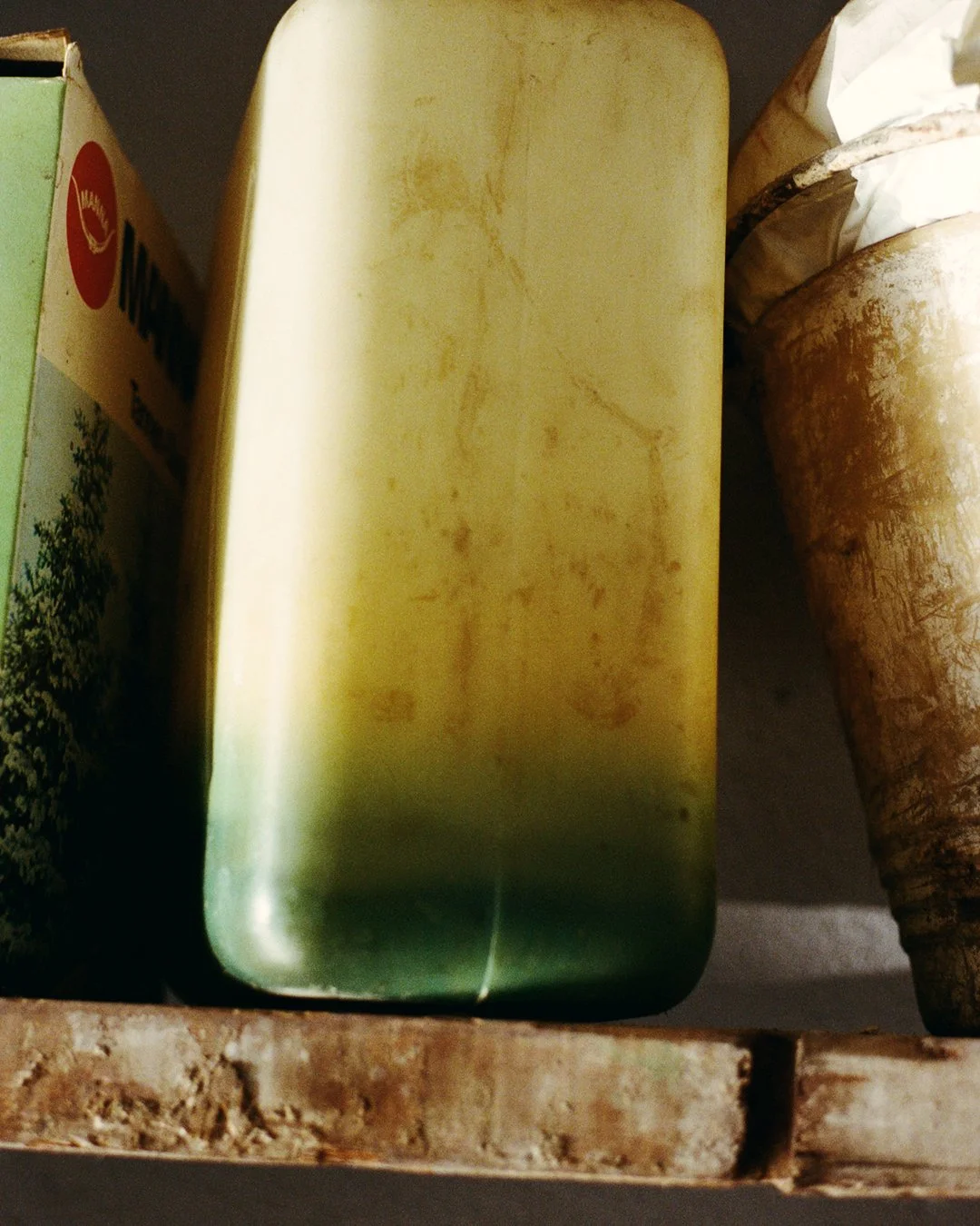 A close-up of an old, yellowed plastic container with dirt or stains on its surface, placed on a worn wooden shelf alongside other items.