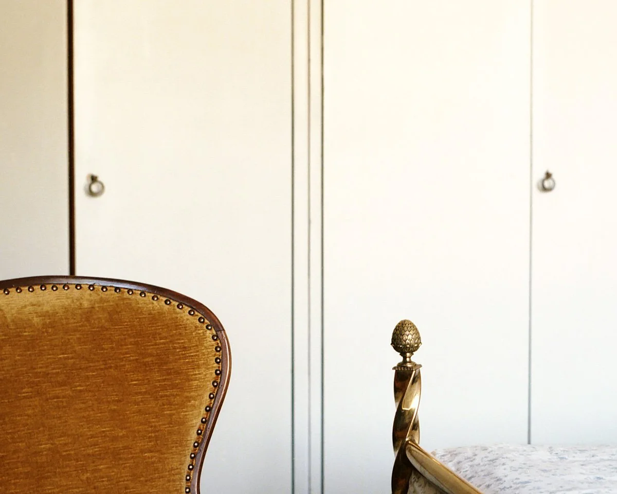 Part of a wooden chair with gold stud accents and a decorative gold bedpost with a pine cone shape on top, against a white closet door.