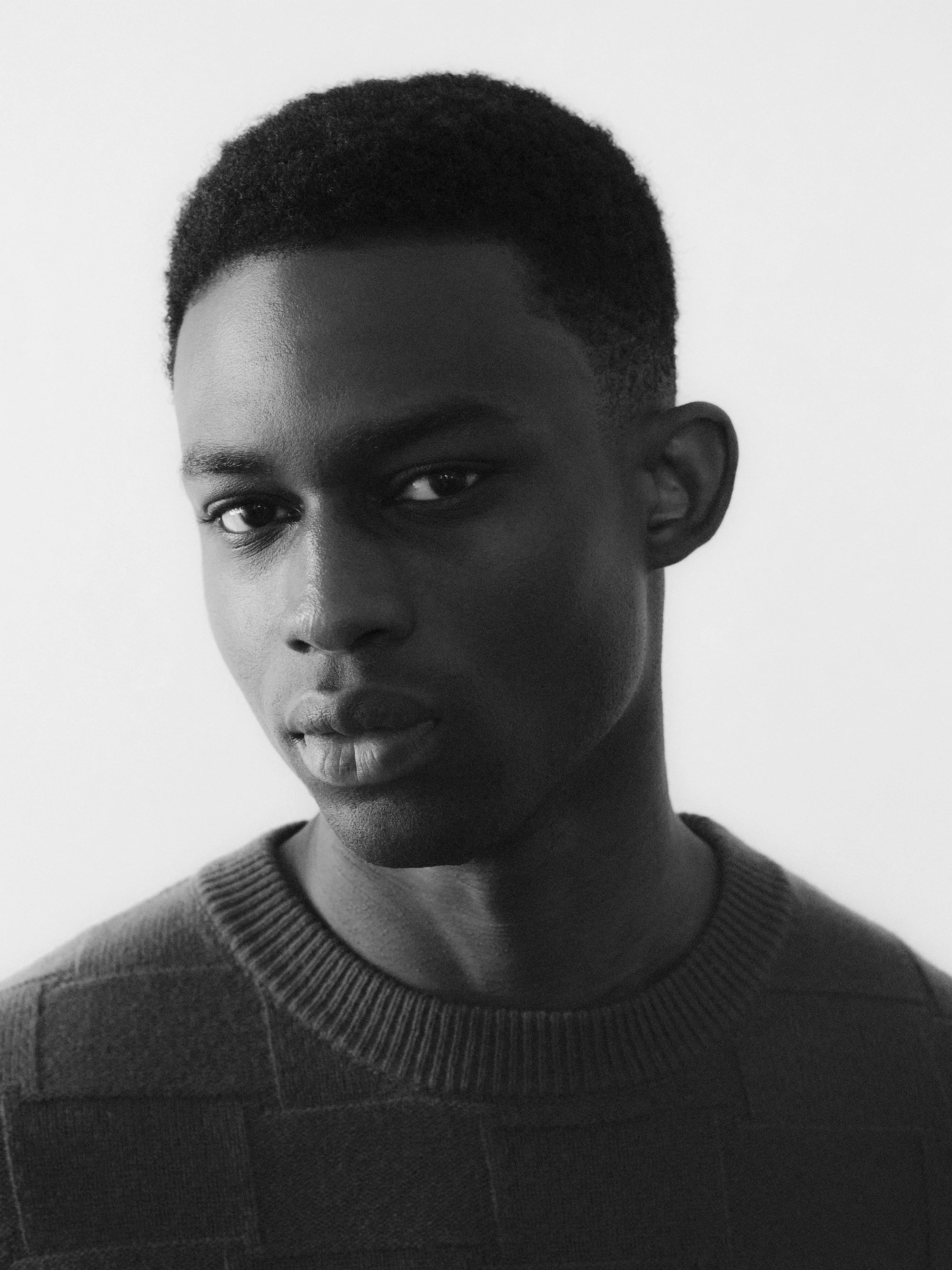 Black and white portrait of a young Black person with short natural hair, wearing a crewneck sweater, looking directly at the camera with a serious expression.