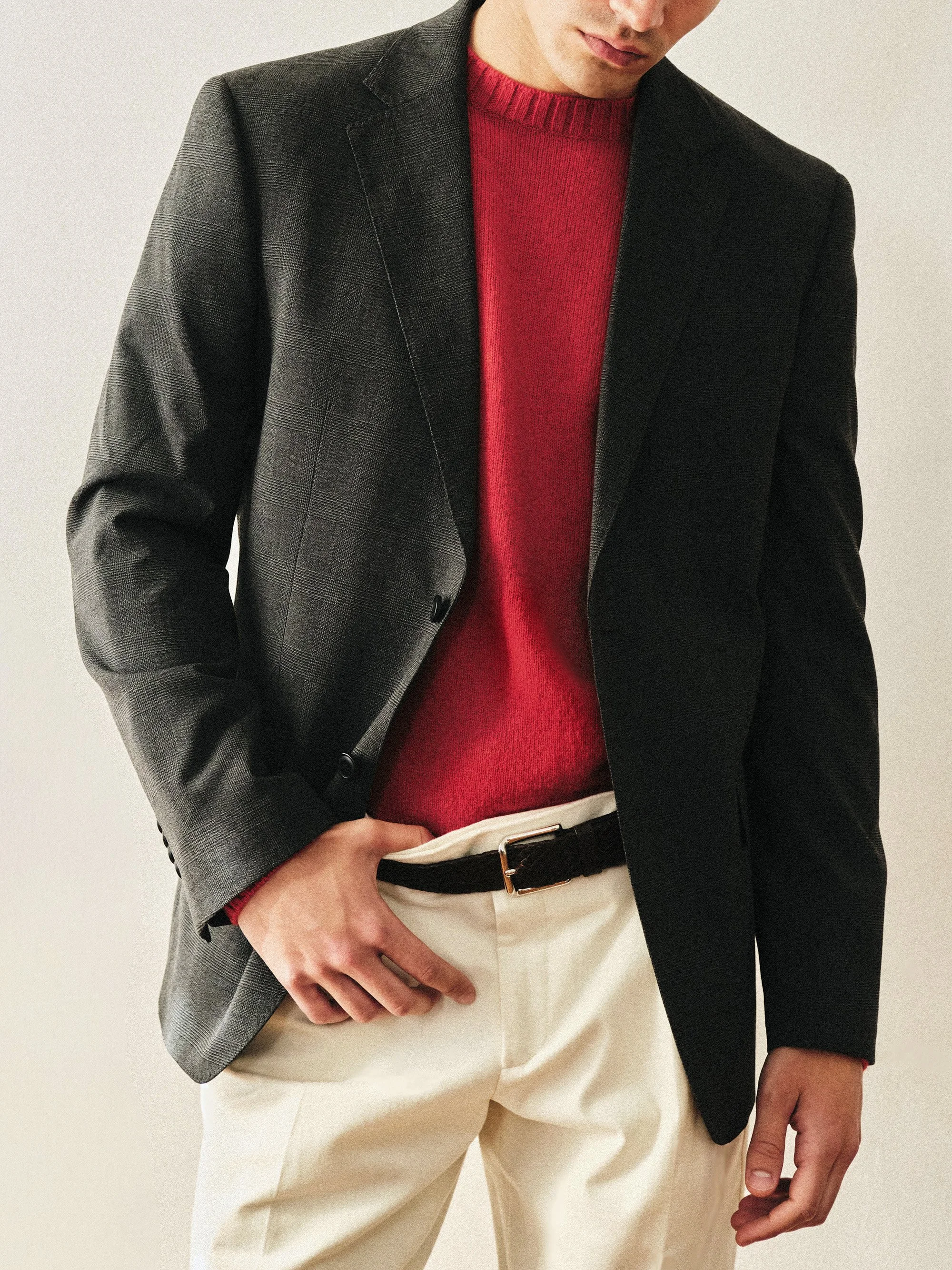 Partial view of a man wearing a dark blazer, red sweater, and beige pants, standing against a plain background.