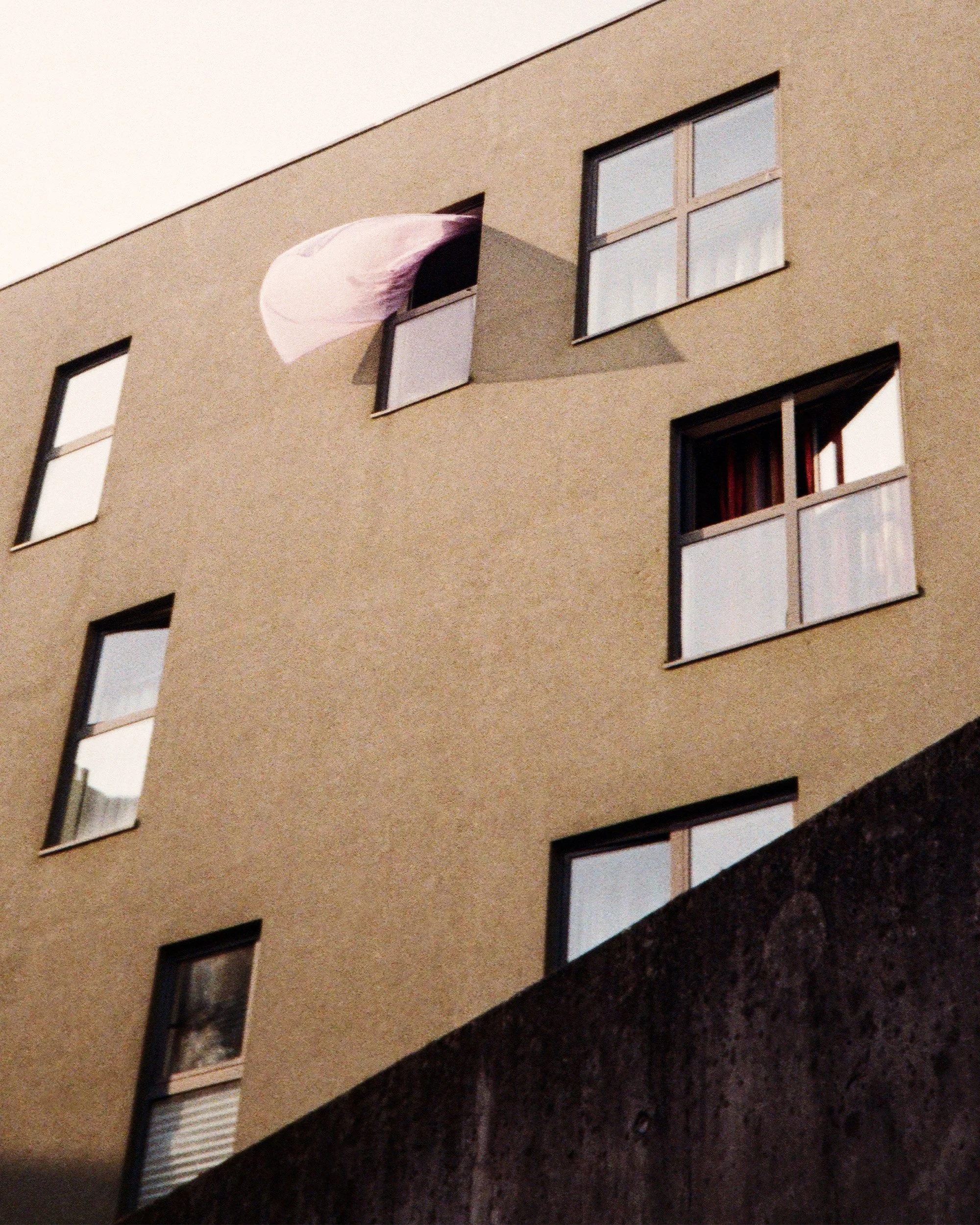 Four windows on the side of a beige building with two open and with a pink and white umbrella hanging out of one window.