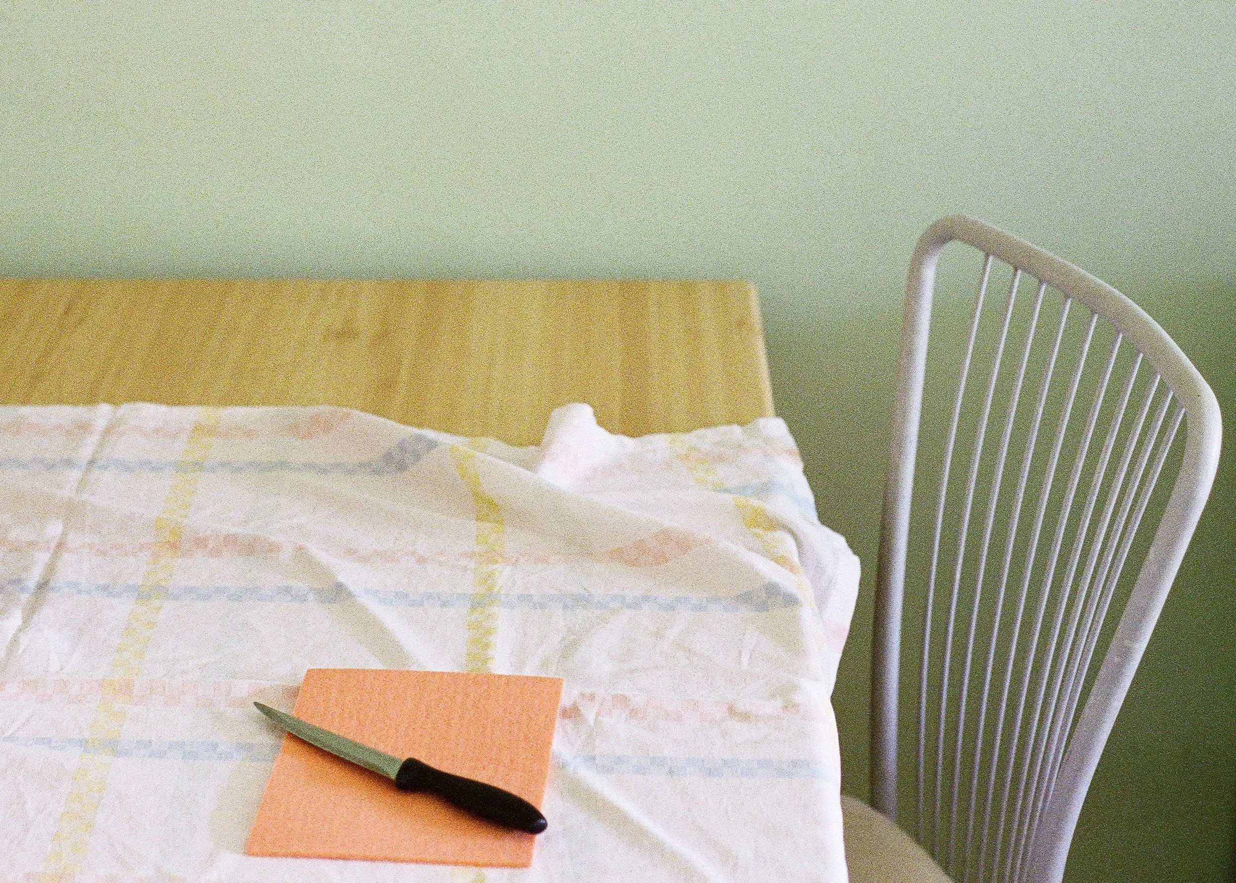 Close-up of a black-handled scissors cutting a small piece of orange paper on a white surface, with a bed or table with a checkered cloth, a white chair, and a green wall in the background.