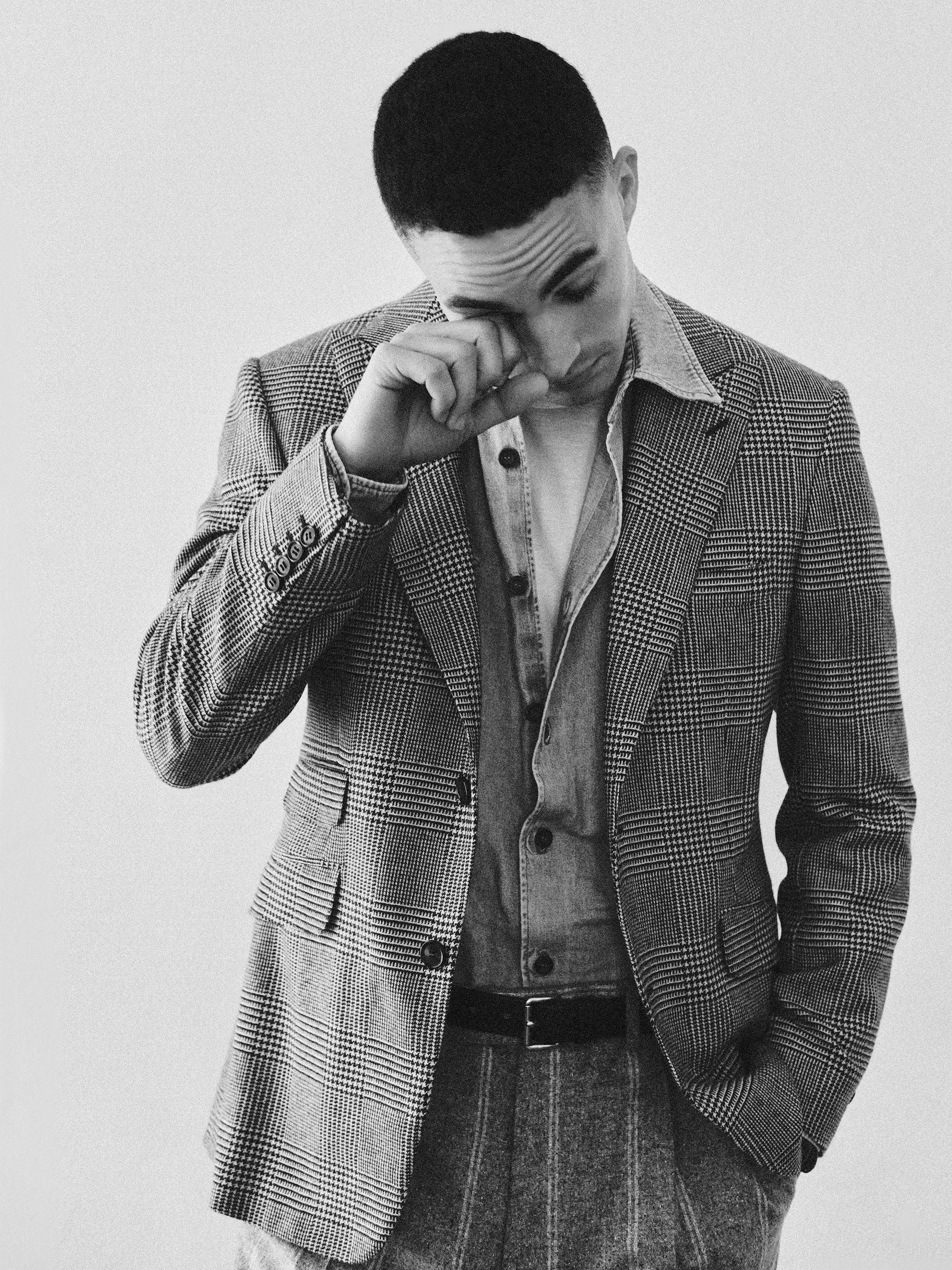 Black and white photo of a young man with short hair, wearing a plaid blazer, denim shirt, and plaid pants, standing against a plain background, with his hand near his face.