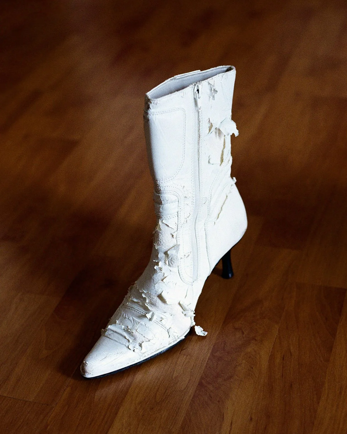 A white high-heeled boot with a pointed toe, partially damaged with peeling material and torn sections, placed on a wooden floor.