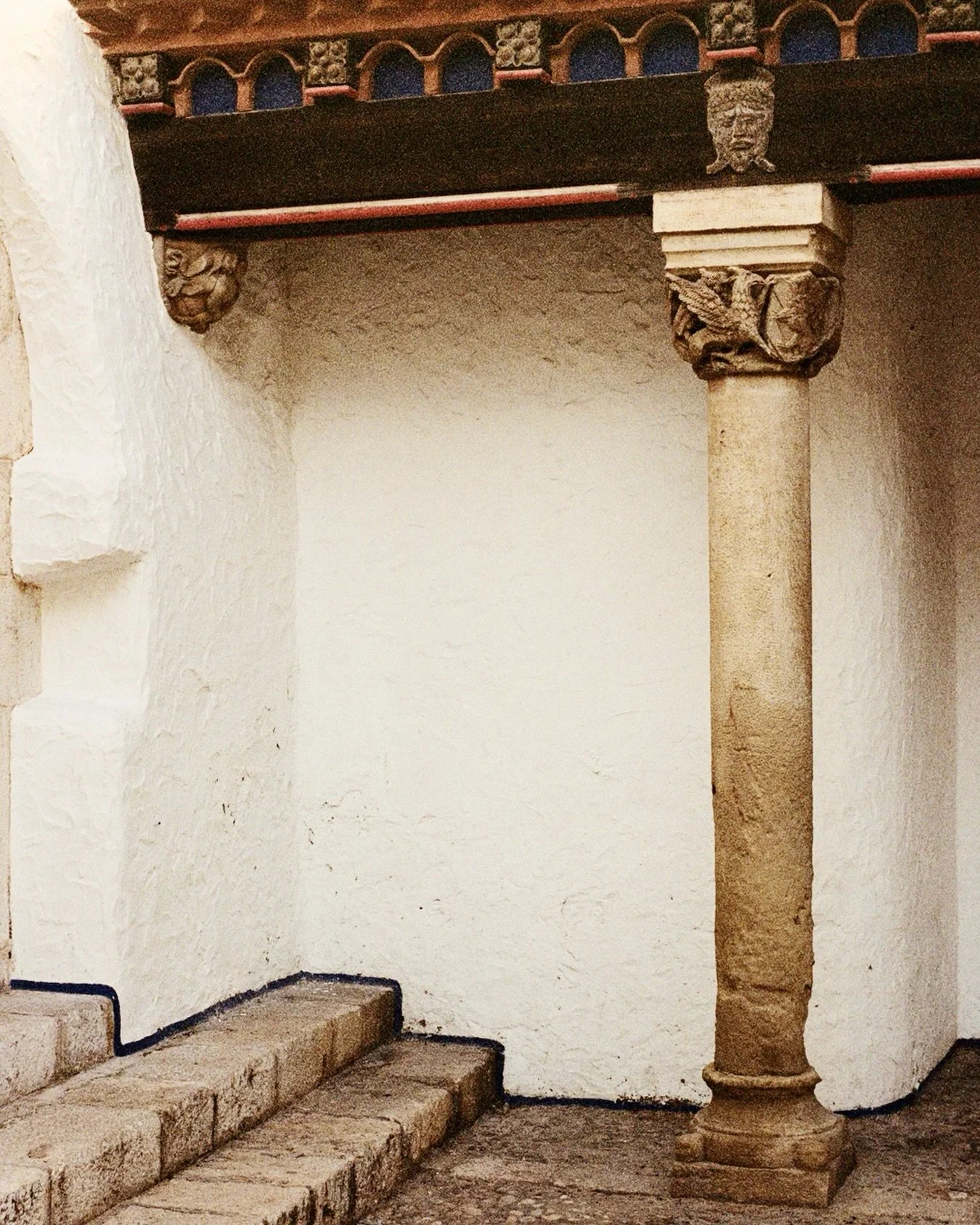 Stone steps leading up to a white wall with an ornate stone column on the right and decorative carvings on the left. The wall has a textured surface, and the steps are old and weathered.